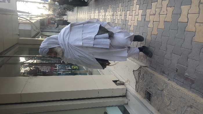 The image depicts a daytime street scene in Quetta City Tehsil, Pakistan. An elderly man is walking on a paved sidewalk, moving from left to right across the frame. He has a white beard and is dressed in a traditional white shalwar kameez, a white turban, and a heavy white shawl draped over his shoulders, partially revealing a dark green garment underneath. He wears black shoes and black socks. On his right side, a small, dark object (possibly a bag or tasbih/prayer beads) hangs from his hand.
To the man's left, a modern storefront with large glass windows reflects trees and the sky. A white pillar separates the windows, and a "50%" discount sign is visible inside the store. The lower section of the pillar has a small, open rectangular recess. The sidewalk directly adjacent to the building is partially damaged, showing exposed concrete and underlying tiling, with a small square drain cover visible. The main sidewalk where the man walks consists of interlocking grey and beige pavers.
In the background, other pedestrians are visible on the sidewalk, along with several parked motorcycles lining the street. Additional buildings, utility poles, and overhead wires are present, indicating an urban setting. The lighting suggests a bright, sunny day.