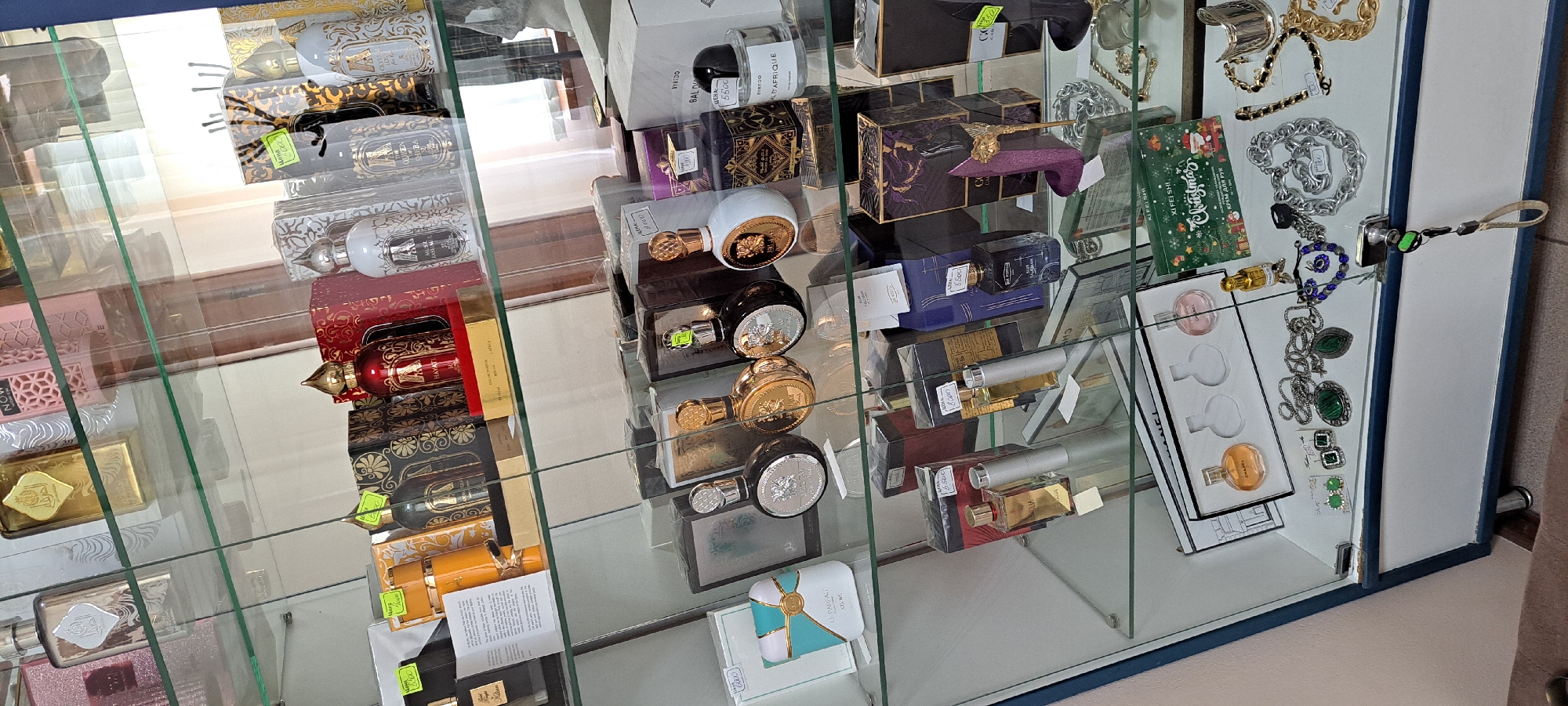 The image shows a display of perfumes and jewelry inside a glass-fronted cabinet. The cabinet is filled with various bottles of perfume, some in boxes, and an assortment of necklaces, earrings, and bracelets. The scene appears to be a retail environment, possibly a perfume shop or a section of a larger store. The lighting is bright, illuminating the products clearly. The background consists of shelves and other display items, suggesting a well-stocked establishment. The overall impression is one of an organized and visually appealing presentation of consumer goods.