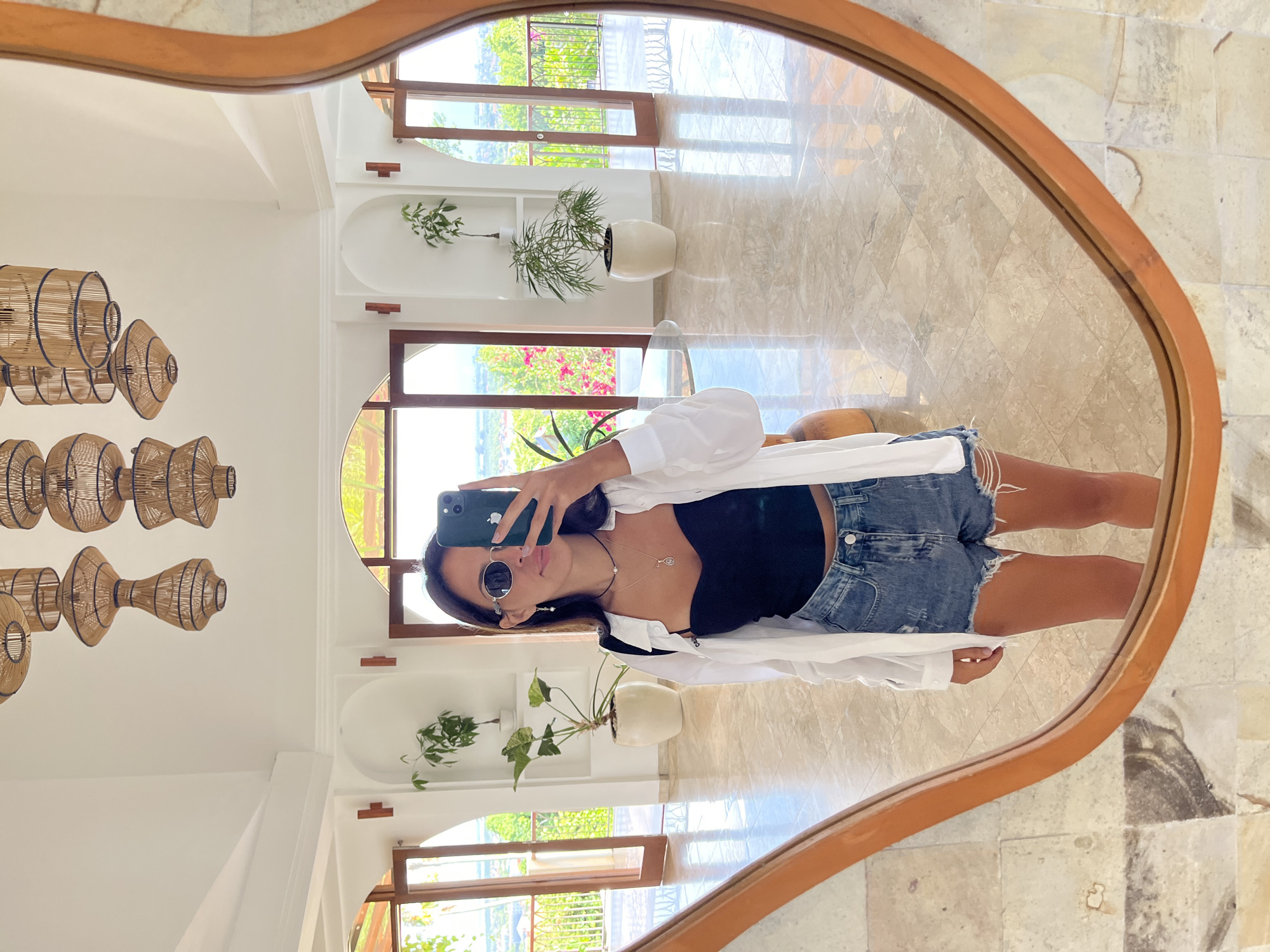 The image is a reflection in a wooden-framed mirror, showcasing a woman taking a selfie. She is wearing sunglasses, a black top, denim shorts, and a white shirt. The reflection includes part of the interior of a building with large windows or doorways that lead to an outdoor area with vegetation. The interior features white walls, rattan light fixtures, plants in pots, and tile flooring. The location is Kuta Selatan, Indonesia.