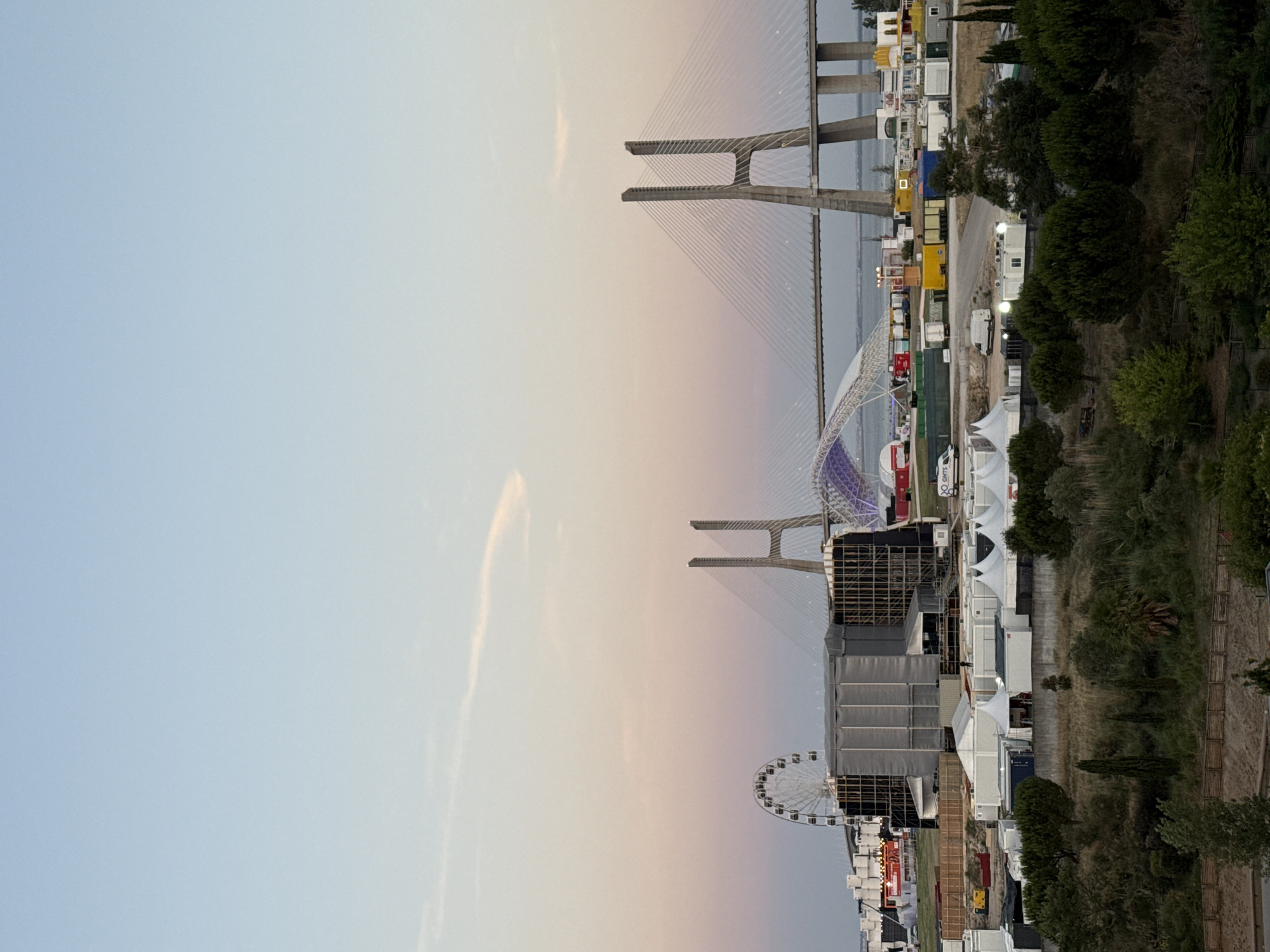 The image shows a landscape with a large cable-stayed bridge spanning a body of water in the background. In the foreground, there is a festival or event setup with numerous white tents, temporary structures, and shipping containers. A Ferris wheel is visible to the left of the bridge. The sky displays a gradient of soft blues and pinks, suggesting either dawn or dusk. This scene is located in Loures, Portugal, specifically at Parque das Nações.