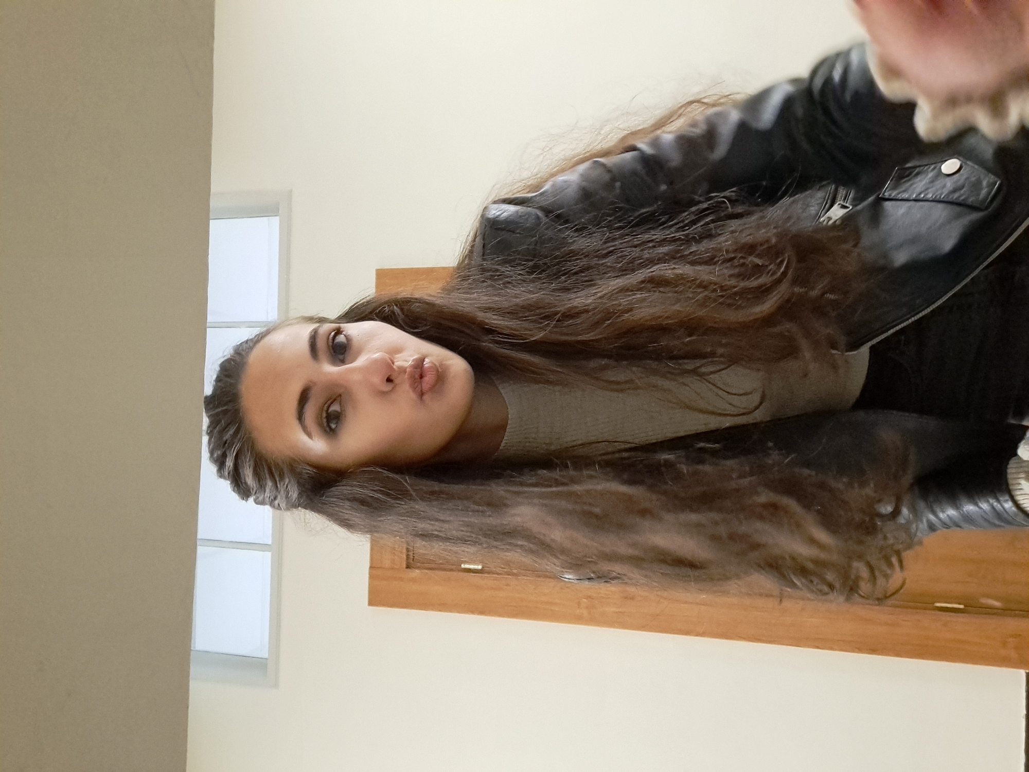 A woman is posing for a selfie. She has long brown hair and is wearing a black leather jacket over a gray top. Her lips are puckered. The background includes a light-colored wall, a wooden door or panel, and a window with white trim. The location cannot be determined from the image.