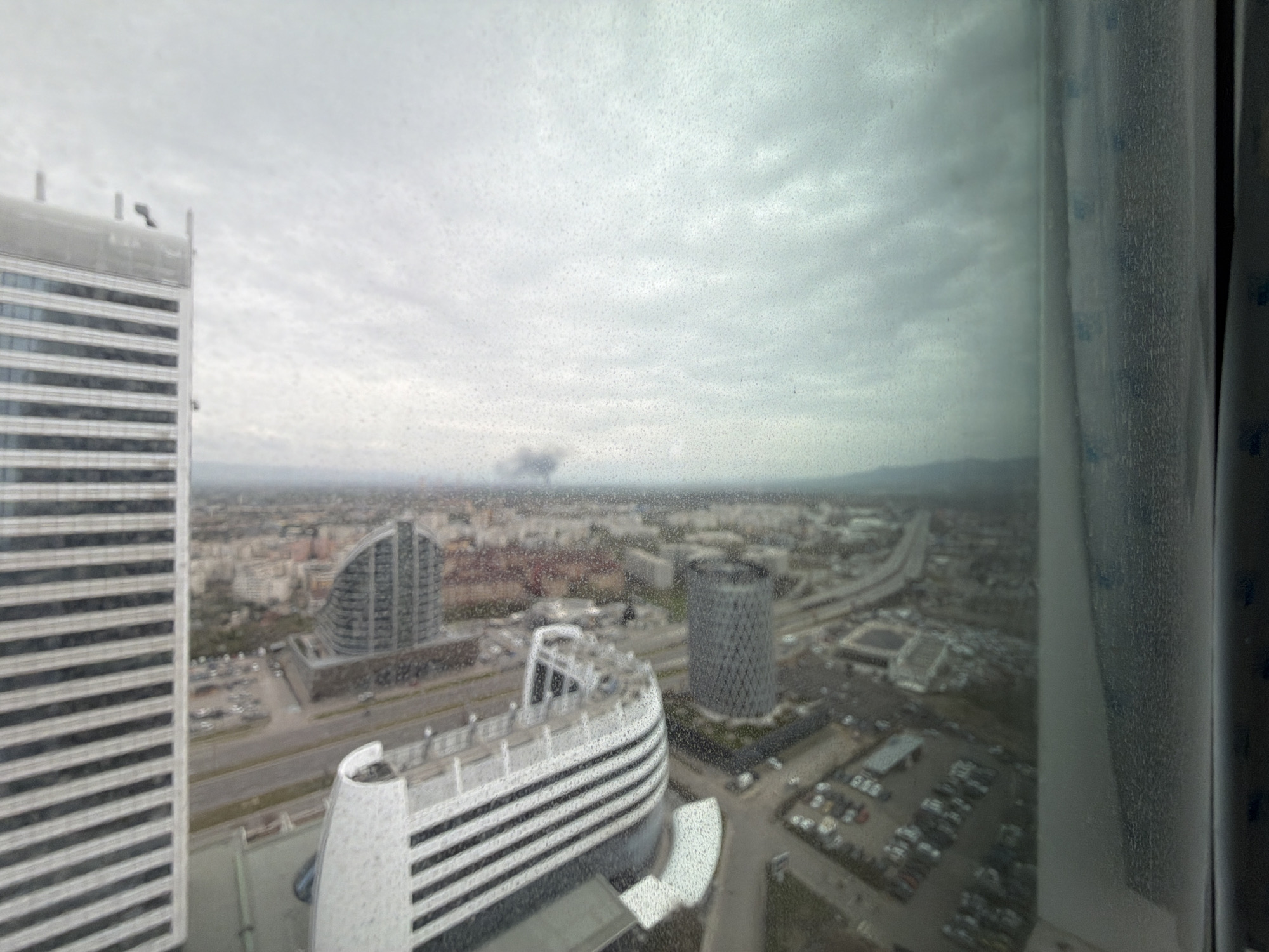 The image presents an aerial view of an urban landscape in Sofia, Bulgaria, observed through a window speckled with raindrops or condensation. The setting is a cloudy, overcast day, lending a muted and grey tone to the scene.
In the immediate left foreground, a tall, rectangular modern building with numerous horizontal window or balcony bands is prominent. Closer to the viewer and slightly to the right, a distinctive white building with a curved, multi-story façade and similar horizontal elements occupies a significant portion of the lower-center. Further back and to the right, a dark, cylindrical building with a textured surface stands out among other city structures. Another modern, angular building with a glass facade is visible in the middle distance.
The city extends into the horizon, characterized by a dense arrangement of buildings, roads, and highways, where indistinct vehicle shapes can be seen. In the far distance, a dark, smoke-like plume or cloud formation is faintly visible against the grey sky. Curtains or blinds frame the right side of the view, slightly obscuring that portion of the city. No people or legible text are visible. The overall impression is of a quiet, somewhat somber urban day.