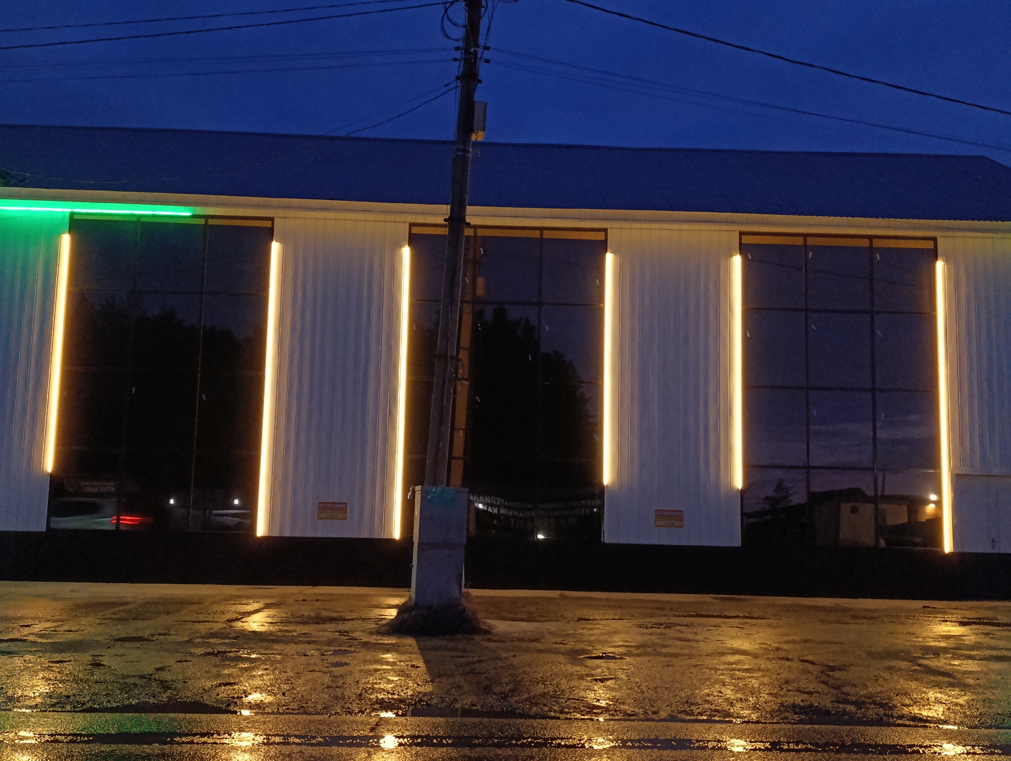This image depicts the illuminated facade of a commercial building in Chaikovsky, Russia, at night. The foreground consists of dark, wet asphalt, reflecting the building's lights in yellow and green hues, indicating recent precipitation. A utility pole with multiple overhead wires stands centrally in the foreground, rooted in a small concrete base. The building features a white, vertically ribbed upper facade over a dark lower section. Three prominent, dark-tinted window sections are visible, each framed by vertical amber LED light strips. A horizontal green LED strip illuminates the upper left portion of the facade. Two yellow warning signs, displaying the Cyrillic text "ОСТОРОЖНО!" (Caution!), are affixed to the white paneling between window sections. A dark blue corrugated roof is visible against a dark blue sky. Reflections in the windows show dark tree silhouettes, faint distant lights, and a partially obscured, inverted banner with Cyrillic text, including "МАГАЗИН" (Store), positioned behind the utility pole. A blurred red vehicle taillight is faintly visible through the leftmost window's reflection. No human subjects are present.