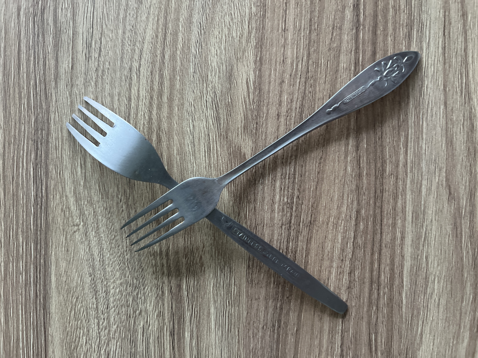 Two stainless steel forks are placed on a wooden surface. The forks are crossed over each other, with the tines of the upper fork pointing upwards and the tines of the lower fork pointing downwards. The surface has a wood grain pattern. There are no people or any other objects visible in the image. The scene does not provide any information about the city of Termez, Uzbekistan.