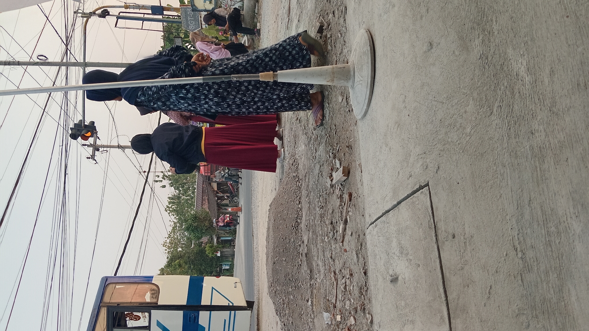 The image depicts a street scene in Trembulrejo, Indonesia, near a traffic light. In the foreground, on the right, a person wearing a dark hijab and a patterned dress stands facing away from the camera. To the left of this person, another individual in a black hooded jacket and a red skirt is also facing away. A light-colored traffic light with the amber signal illuminated is visible in the background, suspended over the road. A white bus with blue accents is parked on the left side of the image. The ground is a mix of concrete and dirt, with a pile of gravel in the middle ground. Several other individuals, some wearing headscarves, are visible in the background, along with motorbikes and a small building. The sky is overcast.