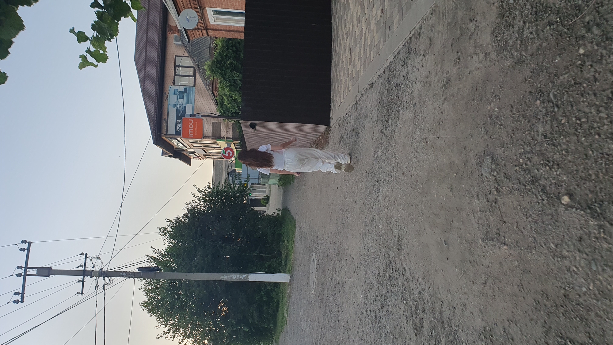 A young woman with long, brown hair, wearing a white t-shirt and loose white pants, walks away from the camera on a gravel path in a suburban area. She is on the right side of the frame, heading towards a paved sidewalk. To her left, a tall utility pole with many wires stands prominently. Behind her, to the right, is a dark, slatted fence bordering a paved area. Further back, a two-story building with a brown roof and brick facade is visible, featuring a sign that reads "КУХНИ" (Kitchens) and another sign with the logo "imou". The sky is clear and light. The scene appears to be in a city, potentially Ust-Labinsk, Russia, given the context.