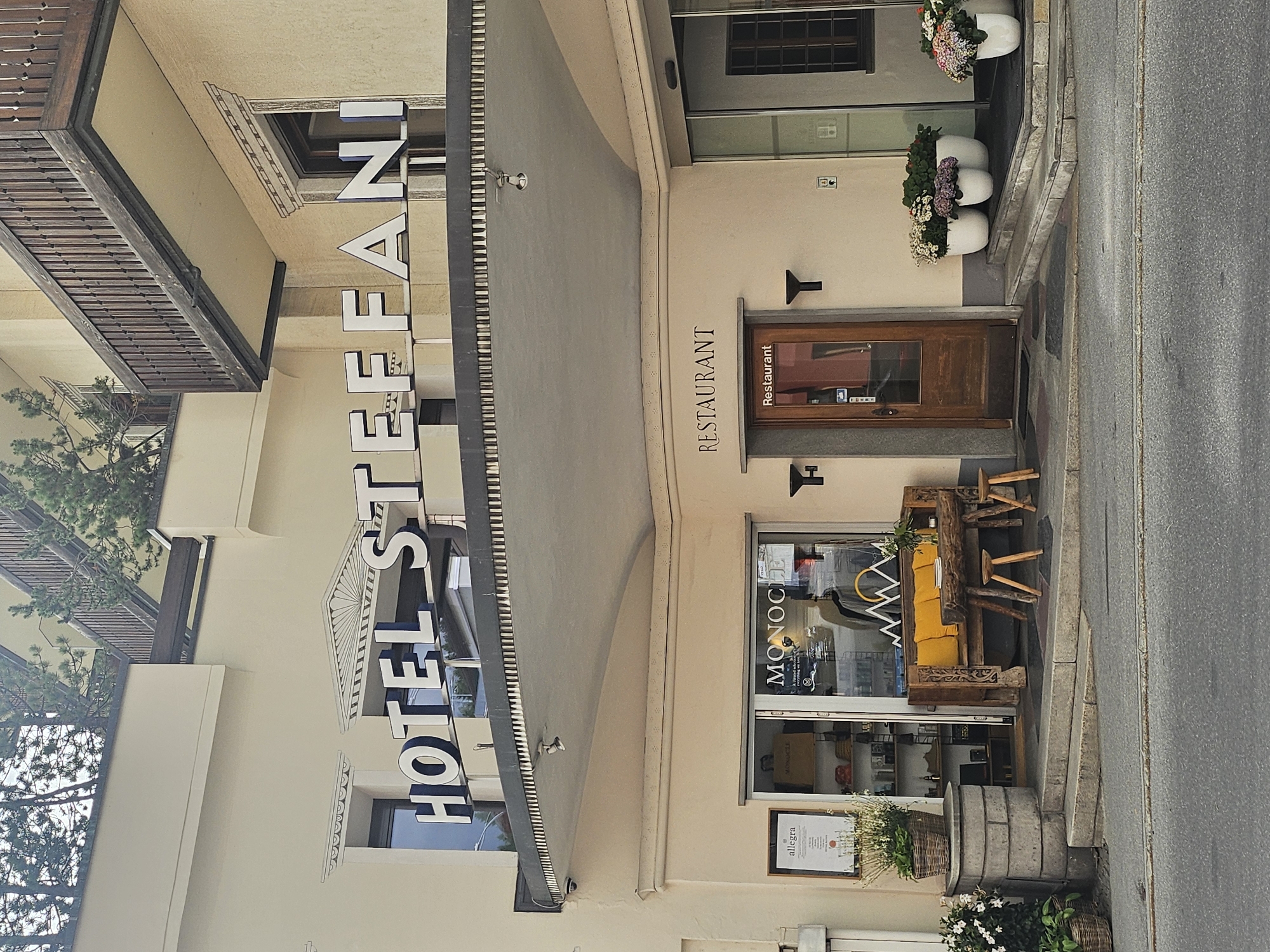 This is a photograph of the exterior of a hotel and restaurant in St. Moritz, Switzerland. The building is light-colored with dark trim and features a prominent sign that reads "HOTEL ST. EFFA NI". The hotel has a traditional facade with awnings and balconies.
In front of the building, there is a street with a sidewalk. The sidewalk features several decorative elements, including planters with flowers, a rustic wooden bench with yellow cushions, and a large barrel planter. A wooden door with a glass panel leads into the establishment, and next to it, a window displays the word "MONOCLE" and a menu. Above the door, the word "RESTAURANT" is visible.
To the left of the hotel entrance, a green tree with dense foliage is growing against the building. The time of day appears to be daytime, with overcast or diffused lighting, suggesting it might be a cloudy day. The overall impression is one of a well-maintained establishment in a European town.
Visible text includes "HOTEL ST. EFFA NI", "RESTAURANT", "MONOCLE", and "allegra".