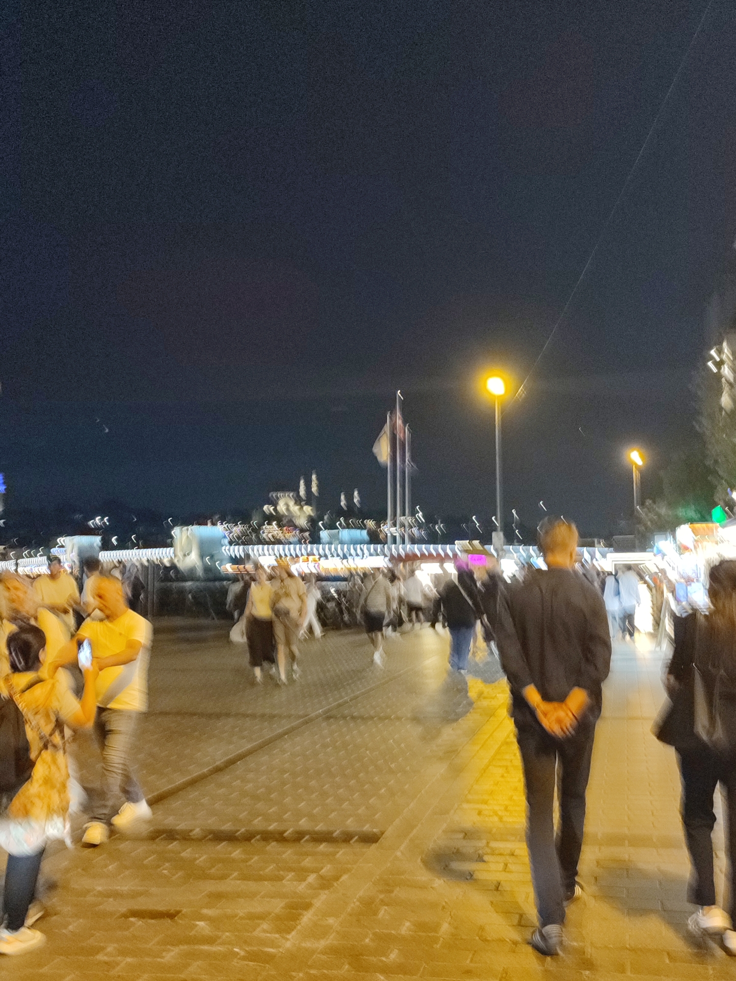 The image depicts a night scene in the city of Beyoğlu, Turkey, with a group of people walking along a cobblestone pathway. The perspective is from behind a person in the foreground wearing a dark long-sleeved shirt and dark pants. In the mid-ground, several individuals are visible, some in motion. To the left, a group of people in lighter clothing are interacting, with one person holding up a mobile phone. The background shows illuminated structures and streetlights, suggesting a bustling urban environment. The overall atmosphere is that of a public promenade at night, with architectural elements and human activity contributing to the scene.