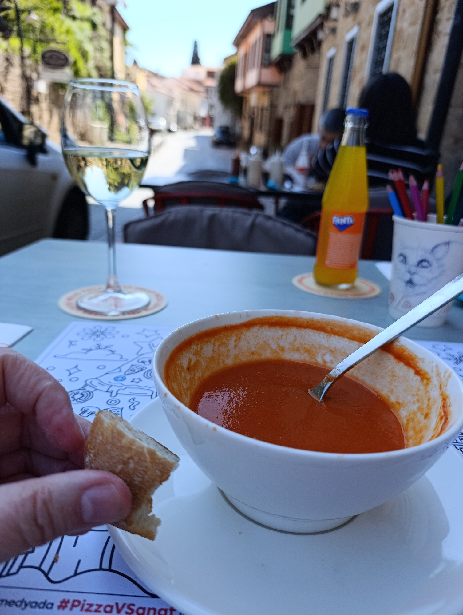 The photo was likely taken at a restaurant in the old town of Antalya, Turkey. A wine glass filled with white wine stands near a bowl of tomato soup with a spoon inside it. A hand holds a piece of bread. Behind the soup, a bottle of Fanta and a cup with colored pencils are visible. The background shows a street with old buildings. Another person is blurred in the background. The table has a placemat with a pizza-themed design and Turkish writing.
