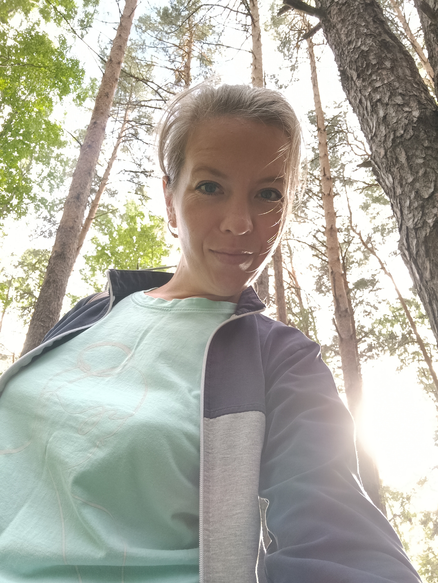 The image is a low-angle selfie of a woman with short, gray hair, looking at the camera with a slight smile. She is wearing a light blue t-shirt with a faint graphic and a gray and navy zippered jacket. The background consists of a forest of tall pine trees with sunlight filtering through the leaves, creating a bright, hazy effect. The scene appears to be an outdoor setting, likely in a park or wooded area within or near the city of Chaikovsky, Russia, given the typical landscape of the region.