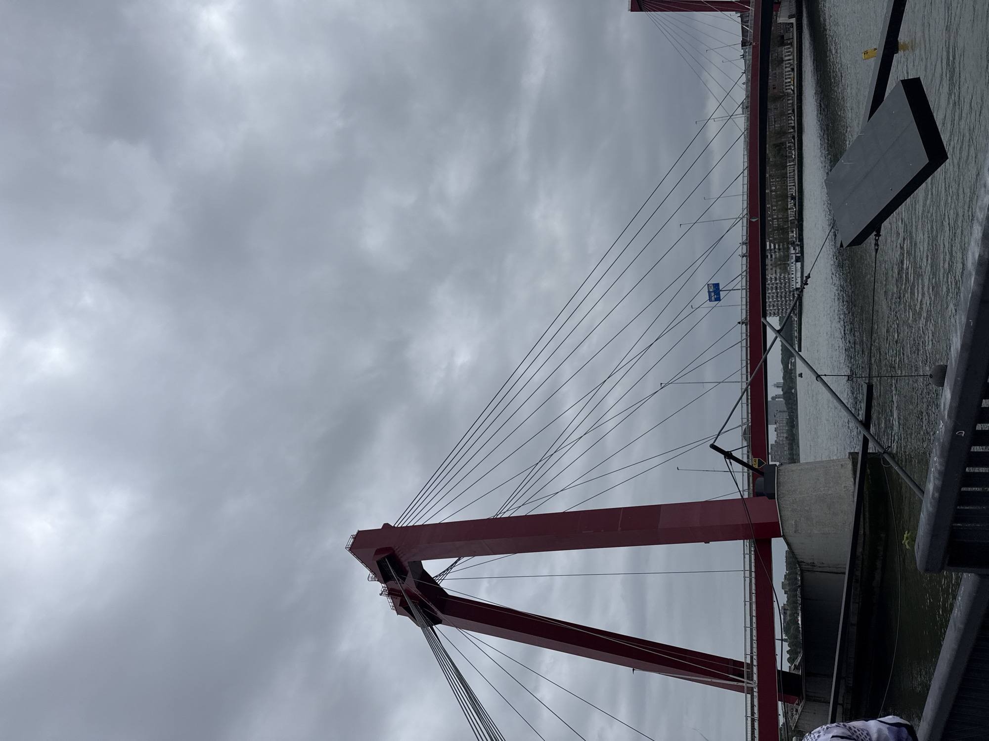 The image shows the Erasmusbrug bridge in Rotterdam, Netherlands, under a cloudy sky. The bridge is a cable-stayed bridge with a distinctive red pylon supporting the main cables. The water below the bridge is calm, reflecting the overcast sky. In the foreground, there is a dark, flat object with some cables attached, which appears to be a piece of debris or equipment. The background shows buildings and other structures along the waterfront, suggesting an urban environment. The overall atmosphere is subdued, likely due to the weather conditions.