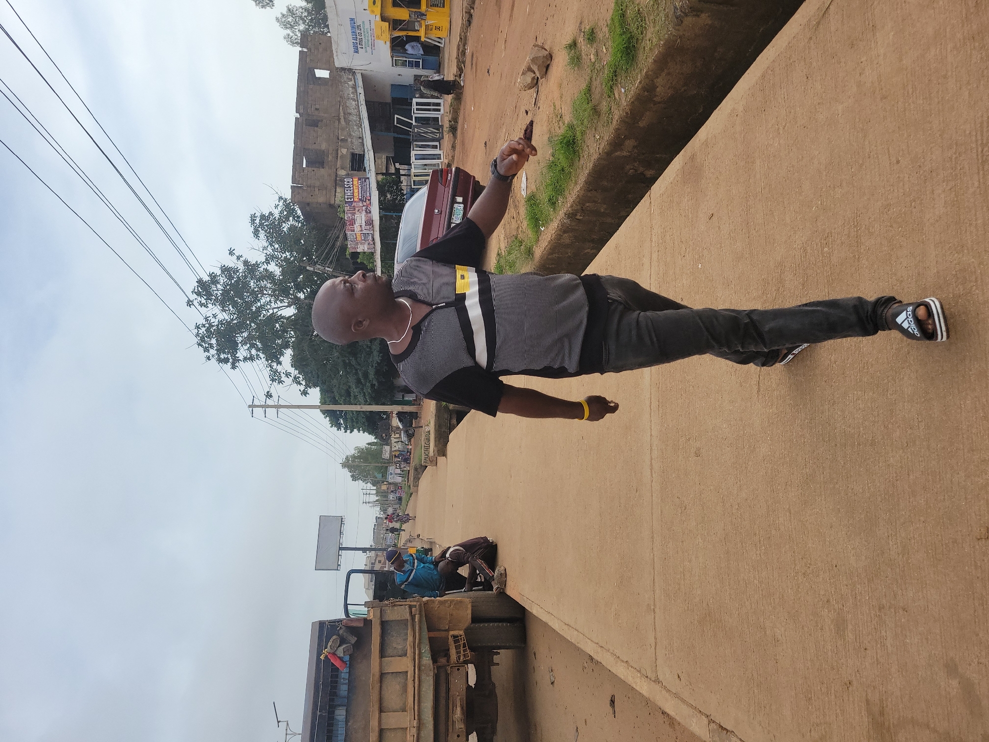 A man walks along a concrete sidewalk in Gura Top, Nigeria. He wears a striped shirt, dark jeans, and sandals. In the background, a truck is parked, and two individuals appear to be working on its tire. Further down the street, buildings line the road, some displaying signage for businesses like "ETHELSCO FURNITURE" and "MARIS ALUMINIUM & STEEL CO. LTD." The sky is overcast.