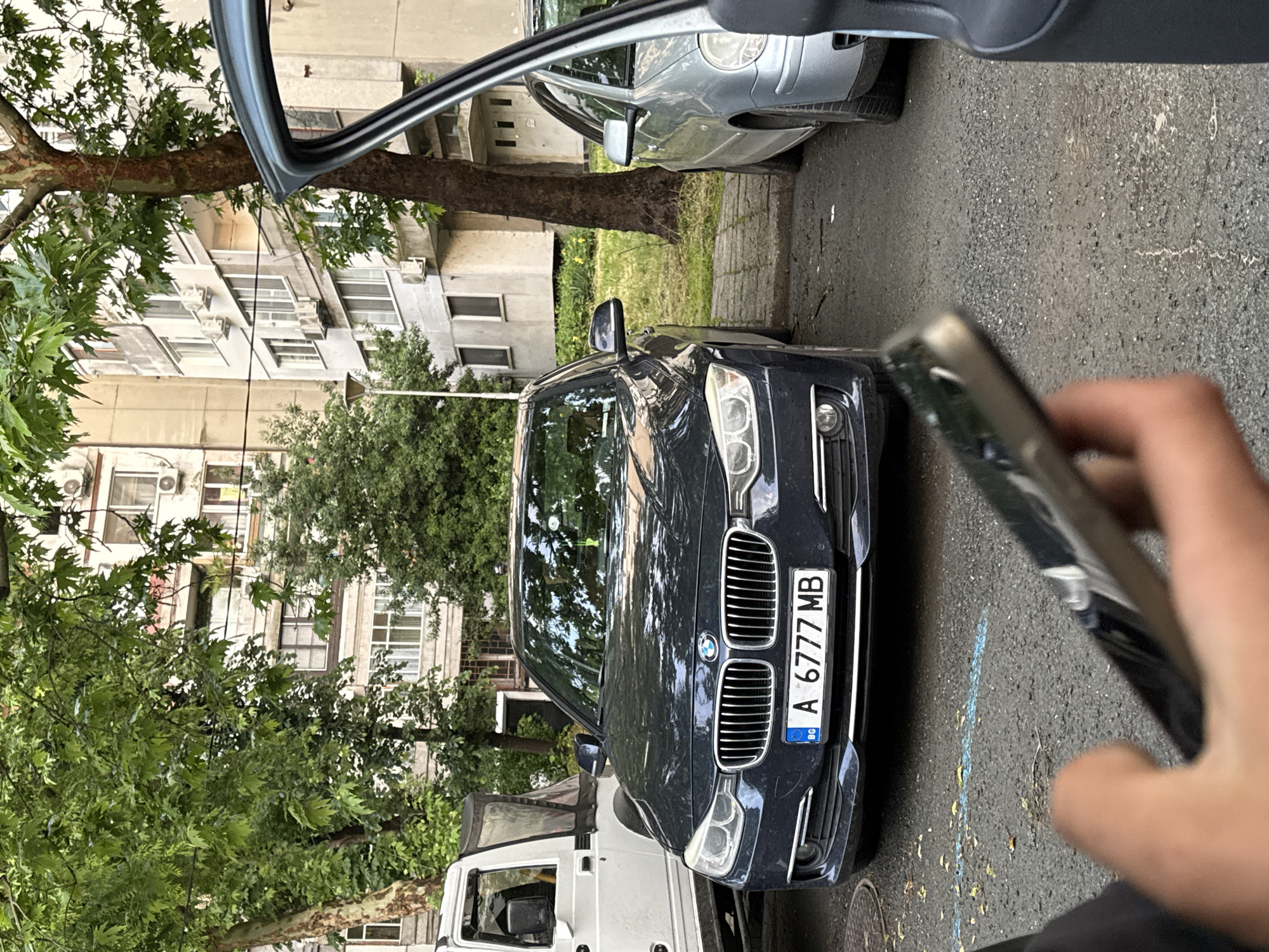 The image shows a dark-colored BMW sedan parked on a street in Burgas, Bulgaria. The car's license plate reads "A 6777 MB" with the Bulgarian flag visible. The car is positioned between a silver vehicle and a white vehicle. An individual is holding a smartphone, possibly taking a picture of the BMW. The street is paved with asphalt. Buildings with multiple windows and air conditioning units are visible in the background, interspersed with trees. The image is taken from inside a car with part of the car's interior visible in the frame.