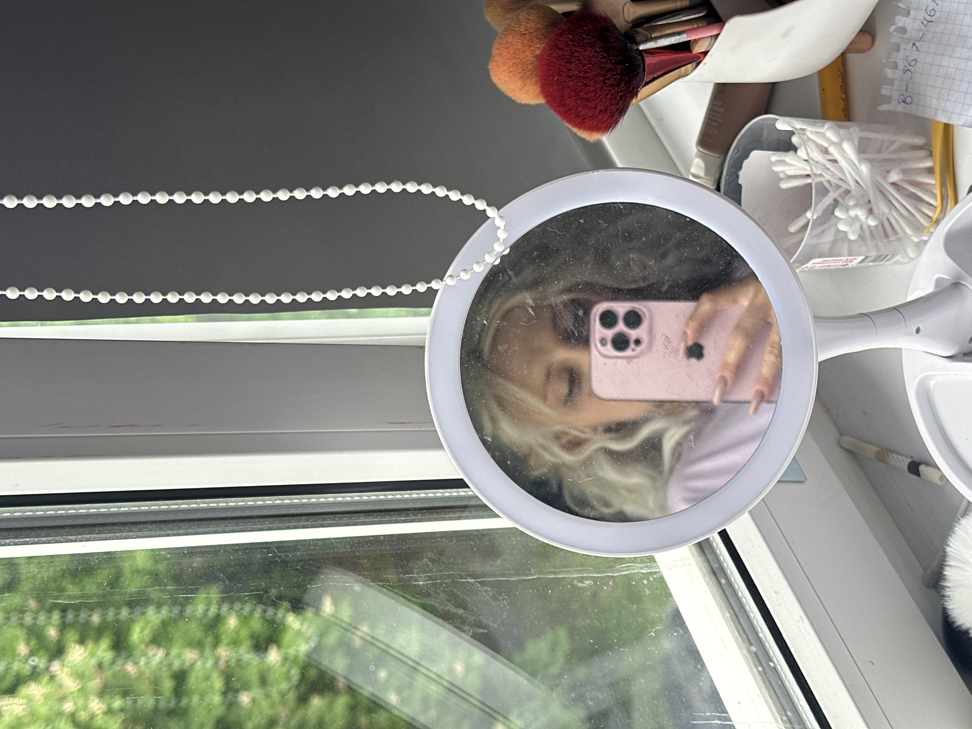 The image shows a close-up of a mirror with a ring light, reflecting a person holding a pink iPhone. The mirror is positioned on a window sill, with a view of greenery outside. To the right of the mirror, there are makeup brushes in a container, a jar of cotton swabs, and a piece of paper with handwritten numbers. A white beaded cord is visible in the upper left corner of the image. The specific location in Stavropol, Russia, cannot be determined from the provided information.