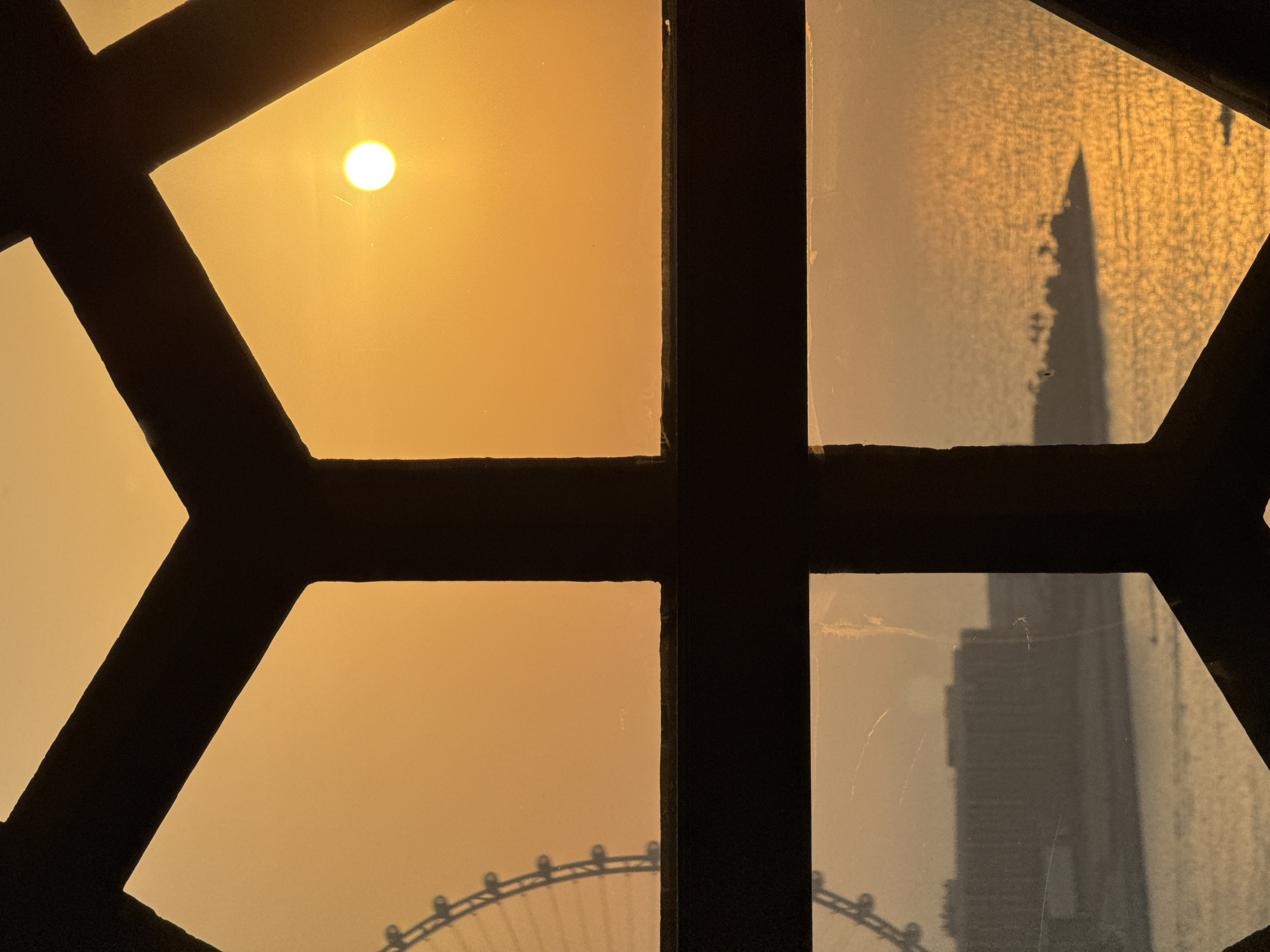 A photograph captures a sunset through a window with a distinctive geometric pattern of dark framing. The sun, a bright, perfect circle, is positioned in the upper left quadrant, casting a warm, golden glow across the sky and the scene beyond. The sky is a uniform, hazy orange, suggesting dust or smog.
In the right half of the image, reflected in the window, a tall, spire-like building casts a distinct shadow, its silhouette sharp against the textured, golden light. Below and to the left, the outline of a large Ferris wheel is visible, its circular structure and spokes silhouetted against the lower part of the sky. The window itself appears slightly grimy, with a few faint scratches visible on the glass, adding a layer of texture to the view.
The overall impression is one of an urban sunset in Dubai, United Arab Emirates. The combination of the sun, the distinctive skyscraper silhouette, and the Ferris wheel are strong indicators of this location. The hazy sky and the warm, diffused light suggest either a dusty environment or the intense heat of the desert sun. The framing of the window creates a sense of looking out from an interior space onto this expansive, atmospheric scene. There is no visible text in the image.