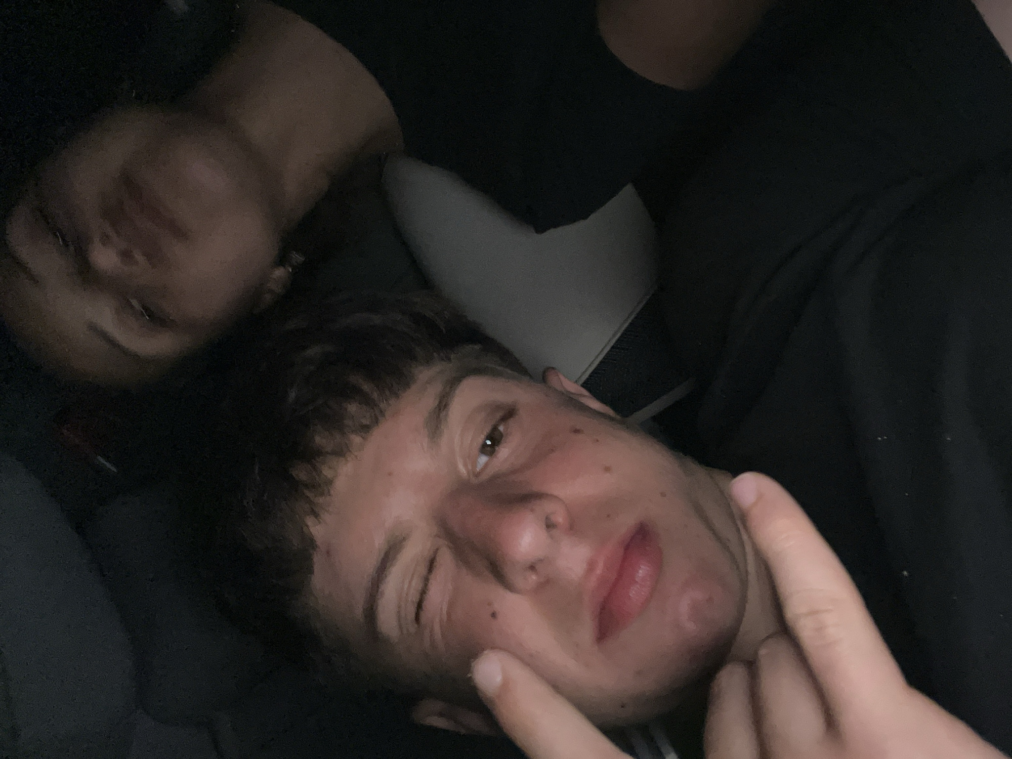 Two individuals are visible. One individual is in the foreground, lying down and winking. Their right hand is raised, with the index finger touching their cheek. The second individual is behind the first, partially obscured and also lying down. The lighting is dim. A light-colored seat is visible in the background. The overall environment appears to be indoors, potentially a car. There is no information in the image to confirm or deny that the location is Popovitsa, Bulgaria.