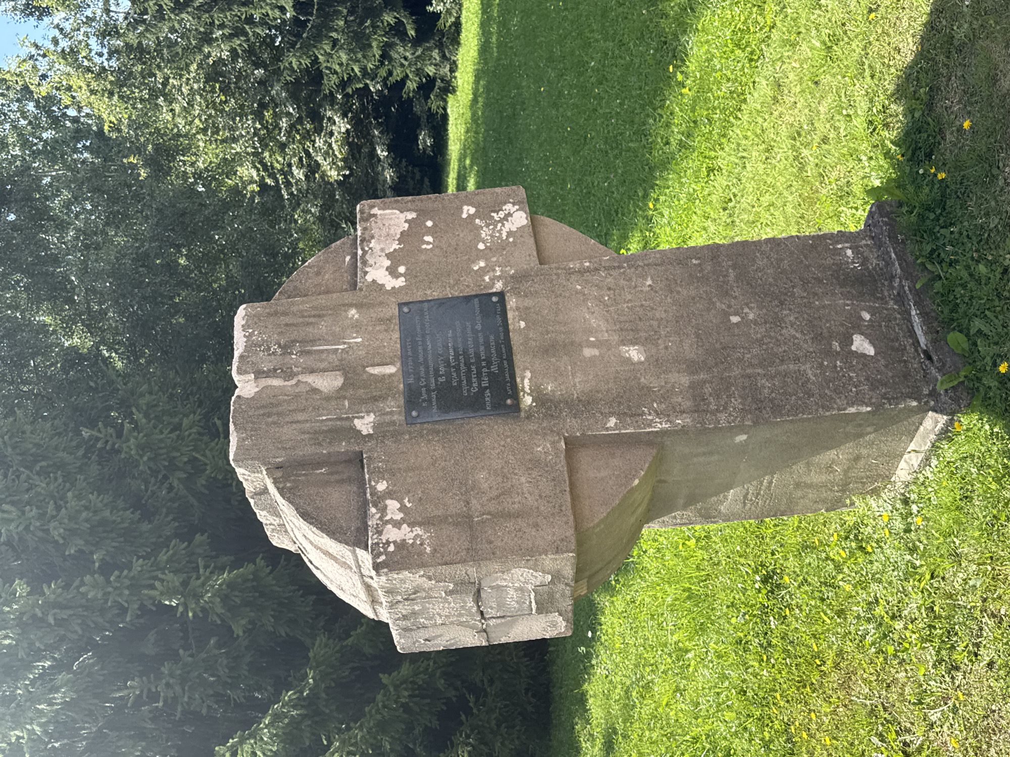 A concrete cross monument stands in a grassy area with trees in the background. A dark plaque is affixed to the front of the cross. The text on the plaque appears to be in Cyrillic script. The monument is weathered, with patches of missing material. The grass is green and appears to be a park or cemetery setting.