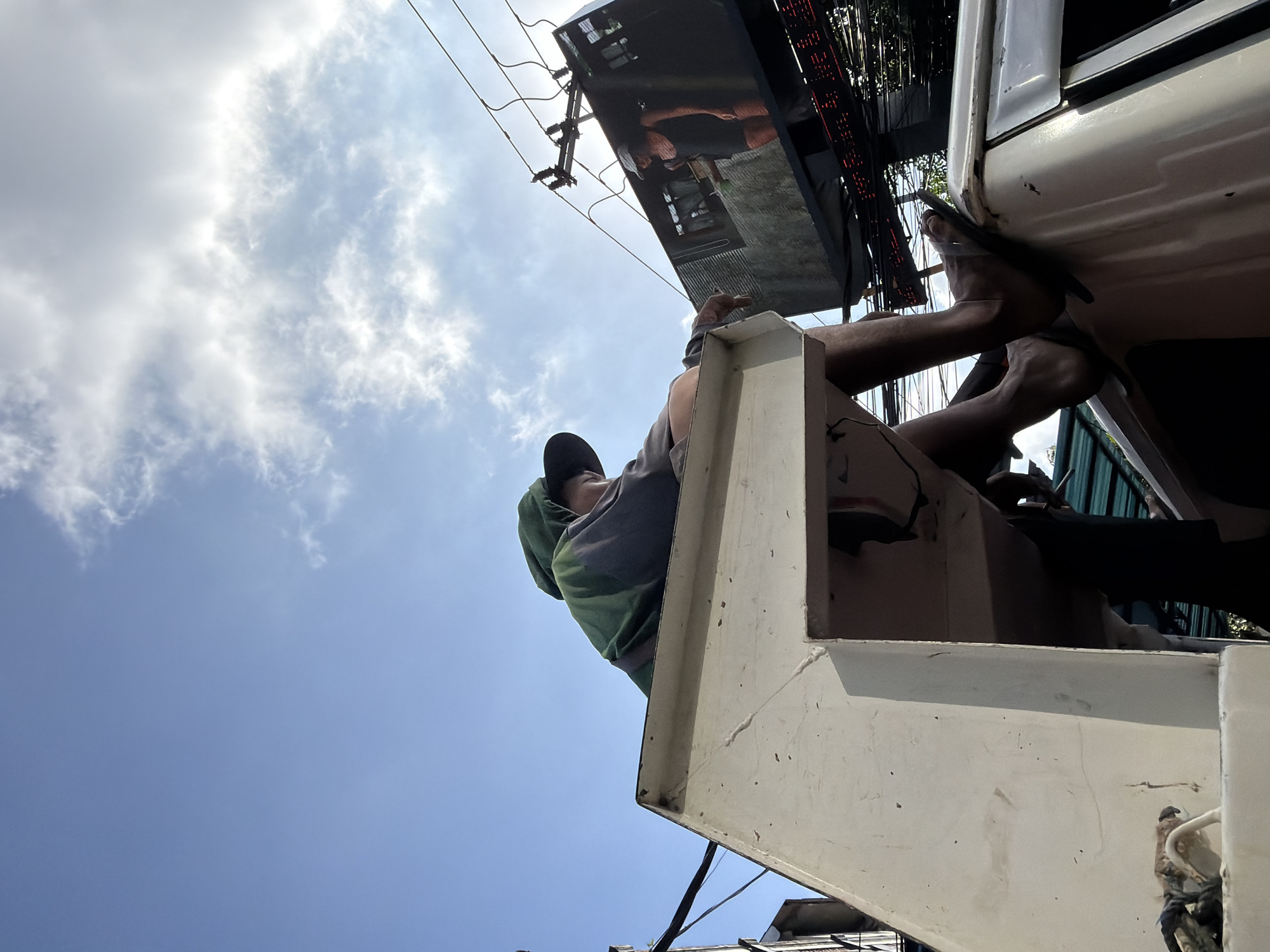 A person is sitting on top of a vehicle, their legs dangling over the side. They are wearing a green hooded shirt and a hat. In the background, a large billboard displays an image of a woman. The scene appears to be in an urban environment with power lines and other electrical infrastructure visible. The sky is blue with some clouds. The context suggests this may be North Kuta, Indonesia.