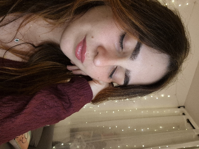 A woman with long brown hair lies with her eyes closed and face in profile. She is wearing a maroon sweater and a delicate necklace. Her lips are glossy. She is in a softly lit room with fairy lights visible in the background. The scene conveys a sense of peace and tranquility.