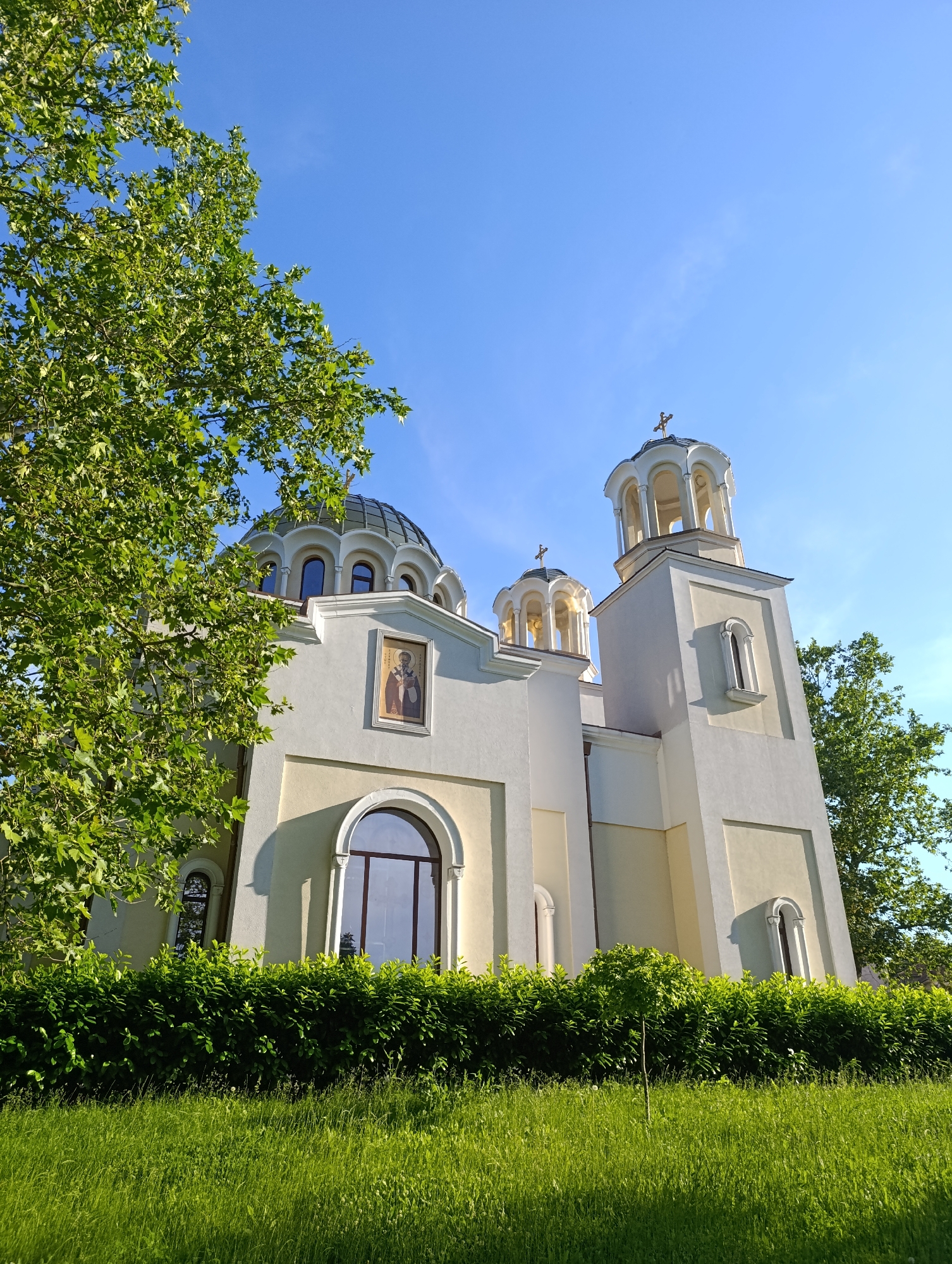 The image shows a bright, sunny day with a clear blue sky. In the foreground, a lush green lawn stretches across the lower portion of the frame, dotted with some taller blades of grass and dandelions. Behind the lawn, a meticulously trimmed hedge forms a verdant barrier.
Dominating the mid-ground and background is a large, white and yellow church with several distinct architectural features. On the left, a large, dark-domed structure with arched windows is partially obscured by the branches of a leafy green tree. To the right of this dome, a tall, slender bell tower rises, topped with a cupola featuring arched openings and a cross. In front of the bell tower, another smaller, similarly designed cupola is visible, also topped with a cross. The church walls are adorned with arched windows and a framed icon of a religious figure.
The scene is bathed in sunlight, casting subtle shadows on the church's facade. The overall impression is one of tranquility and architectural beauty set within a well-maintained natural environment. This church is identified as the "Church of St. Nicholas the Wonderworker" located in the city of Razgrad, Bulgaria.