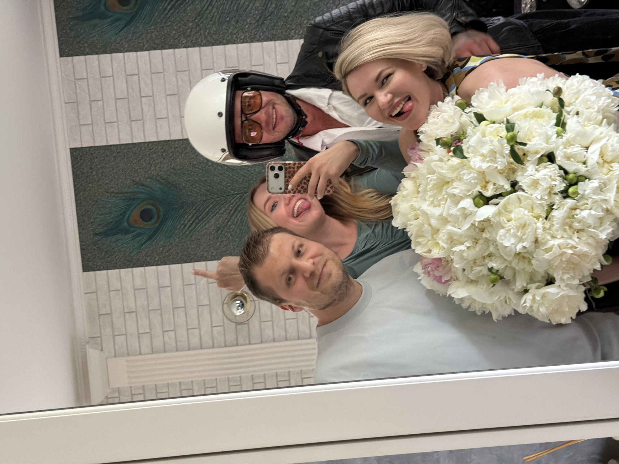 A group of four people are taking a mirror selfie. A man in the foreground wearing a light blue t-shirt is smiling at the camera. To his right, a woman in a green top is sticking out her tongue. Behind them, a man wearing a white motorcycle helmet, sunglasses, and a black leather jacket is smiling. Another woman, with blonde hair, is in front of him, also sticking out her tongue. She is holding a large bouquet of white peonies. The background appears to be a wall with a brick pattern and peacock feather designs. The reflection shows a white pillar and molding, suggesting an indoor setting, possibly a home or a studio. There are no visible landmarks or indicators that would definitively place this scene in Riga, Latvia.