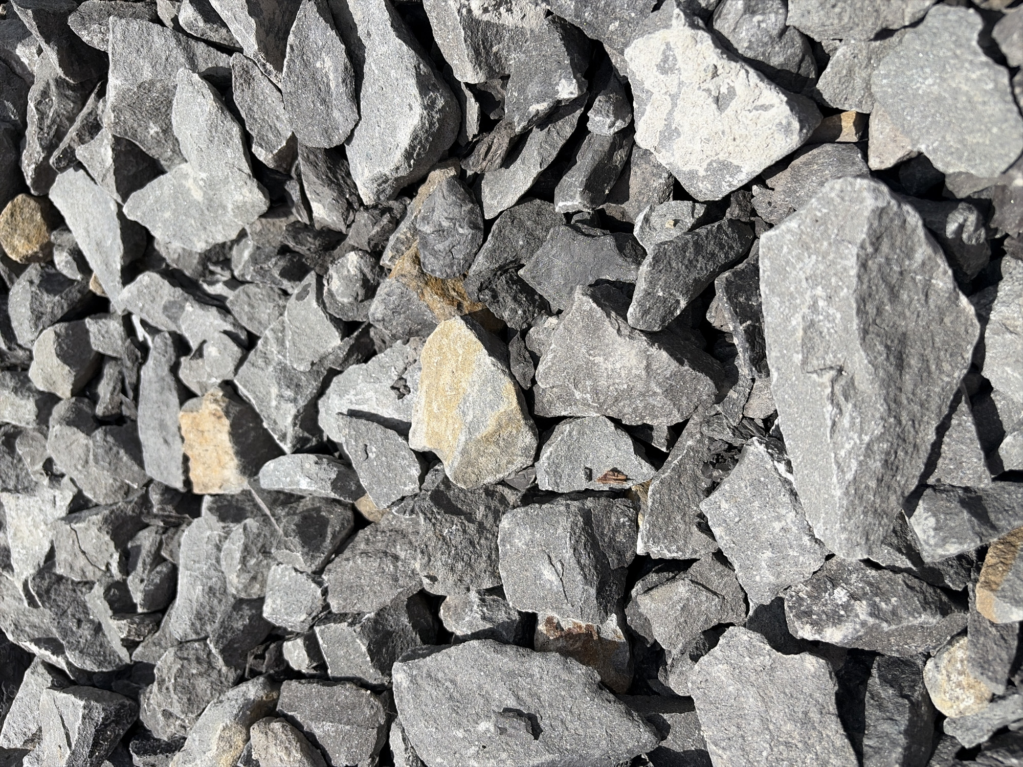 This is a close-up, overhead, static shot of a pile of grey, rough-edged gravel. The gravel consists of angular, irregularly shaped rocks of varying sizes, with a few lighter-colored, tan rocks interspersed within the predominantly grey mass. The lighting suggests a bright, possibly sunny environment, casting shadows that emphasize the texture and three-dimensionality of the stones. The scene is devoid of any human presence, activity, or discernible urban or natural environment beyond the gravel itself. The image does not depict Janowice, Poland or any identifiable geographical location.