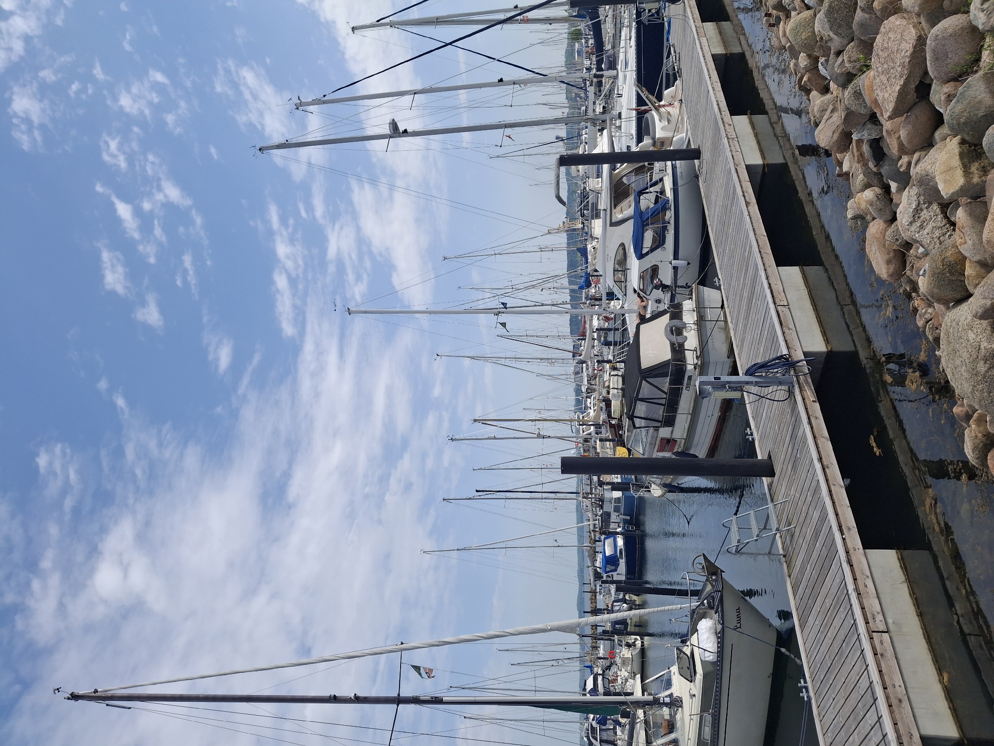 The image depicts a marina in Aabenraa, Denmark. The scene shows a row of sailboats moored side-by-side along a wooden pier. The masts of the sailboats extend high into a sky with scattered clouds. To the right of the pier, there is a rocky embankment, where the land meets the water. The focus is on the boats and the marine environment, with the sky occupying a significant portion of the frame. There are no visible people in the immediate view, although some human presence is indicated by the boats and their arrangement.