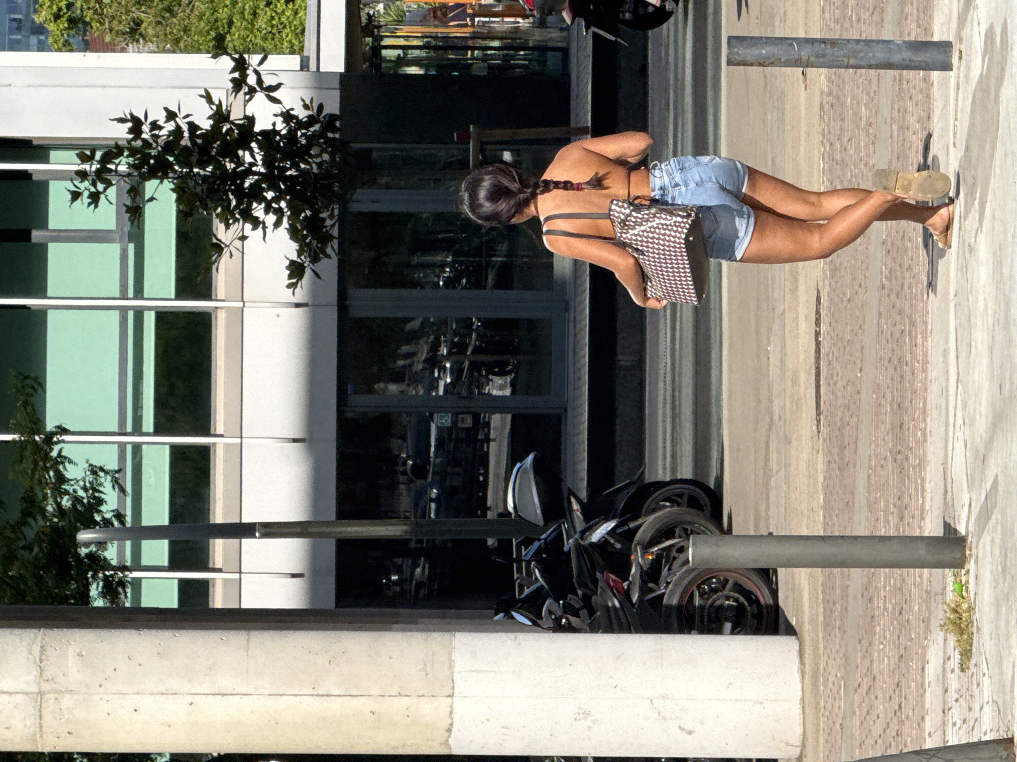A woman with dark hair in a braid is walking on a sidewalk in L'Hospitalet de Llobregat, Spain. She is wearing denim shorts and a halter-style top, and carrying a patterned tote bag. Behind her, several motorcycles are parked. The background features a modern building with large green-tinted windows and a concrete column. The ground is paved with cobblestones and concrete. Sunlight casts shadows on the ground.