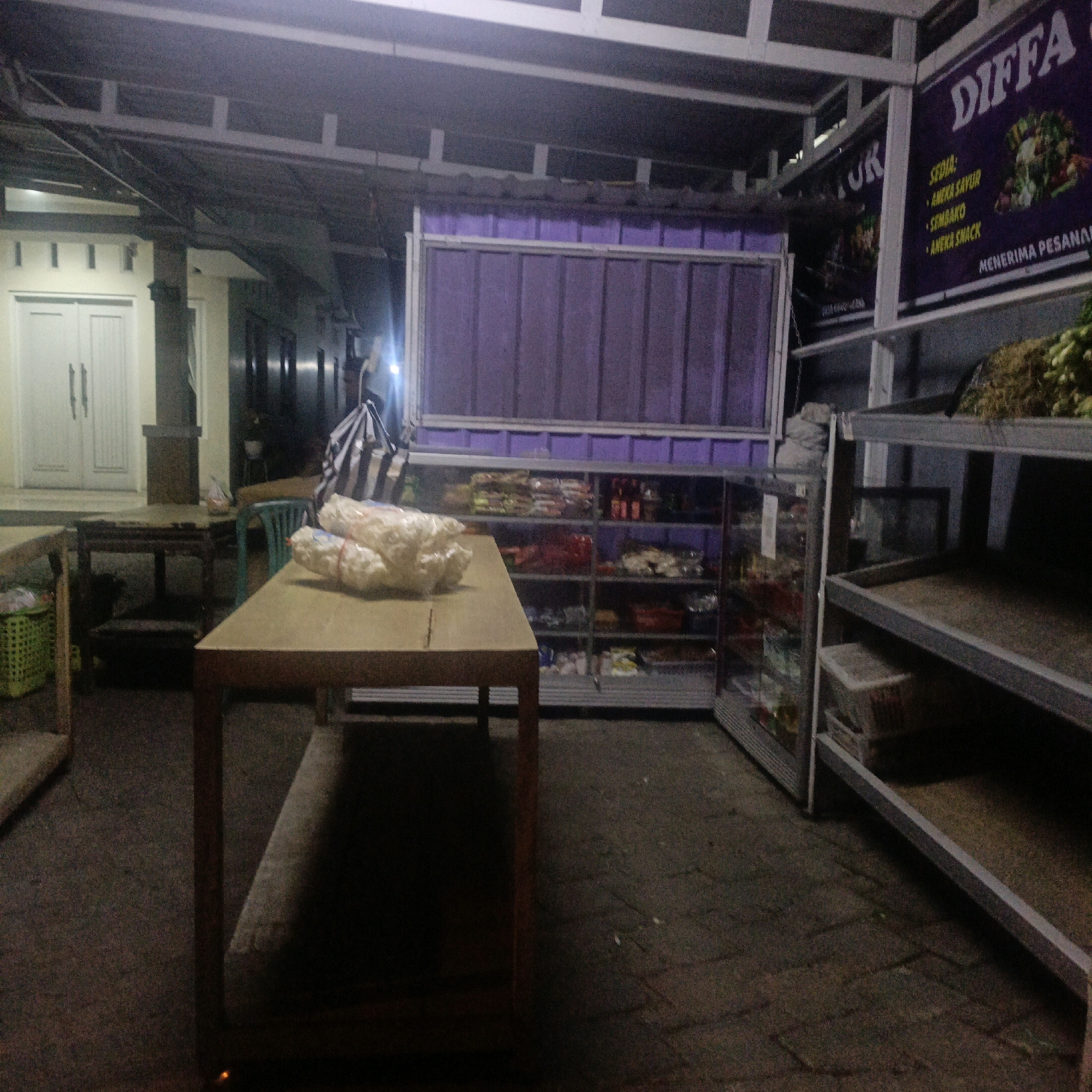 The image depicts a street-side stall in Salatiga, Indonesia, likely operating in the evening or at night due to the lighting conditions. The stall appears to be selling food items, with shelves stocked with various packaged goods, some of which are visible in a glass display case to the right. A prominent sign with the text "DIFA" is visible, along with other text that is difficult to discern but appears to be in Indonesian, listing items such as "ANEKA SAYUR" (various vegetables), "SEMBAKO" (groceries), and "ANEKA SNACK" (various snacks).
In the foreground, a wooden table is positioned, with several large bags of what appear to be snacks or crackers on top. The table is simple, with wooden legs and a flat top. In the background, a building with white double doors and some decorative elements is visible. The stall structure itself seems to be made of metal and possibly corrugated plastic, with a purple panel in the middle section. The ground is paved with what looks like interlocking bricks. To the left, a green plastic basket and a wooden table are also present. The overall scene suggests a modest retail operation, typical of small businesses in Indonesian cities. There are no people clearly visible in the image.