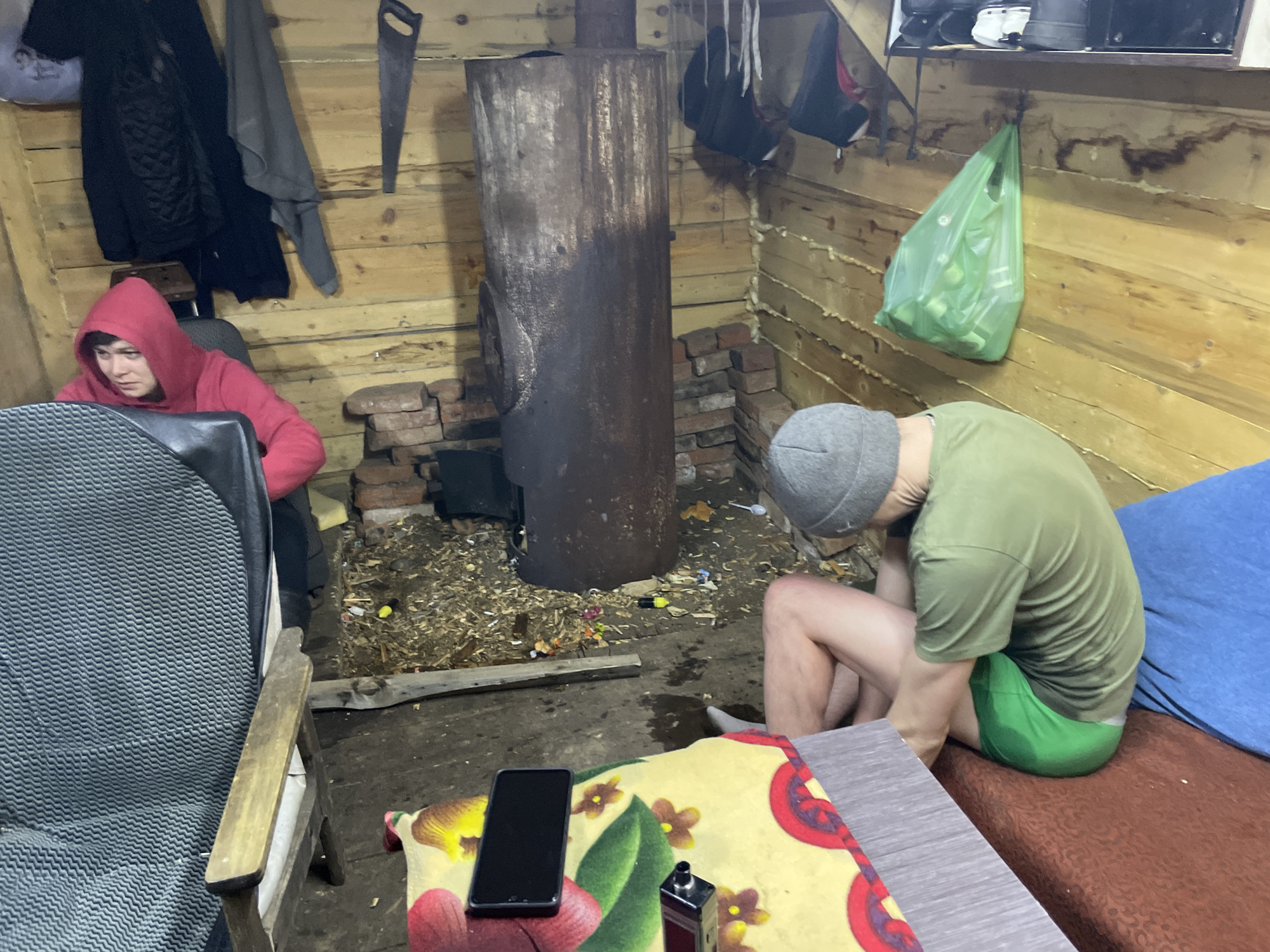 The image depicts an interior scene, possibly a rustic cabin or outbuilding. The walls are constructed of wood planks. A rusted metal stove stands in the center-left, surrounded by bricks and debris on the floor. To the left, a person in a red hooded sweatshirt sits in a chair, appearing to be engaged with something out of frame. To the right, another person, wearing a gray hat and green shirt, sits on a bed covered with a blue sheet. A table covered with a floral-patterned cloth is in the foreground, and a smartphone and other objects are placed on top. A plastic bag hangs from the wall above the person on the bed. It's difficult to determine the city of Nizhnii Bugrysh, Russia as a location from the information provided in the image.