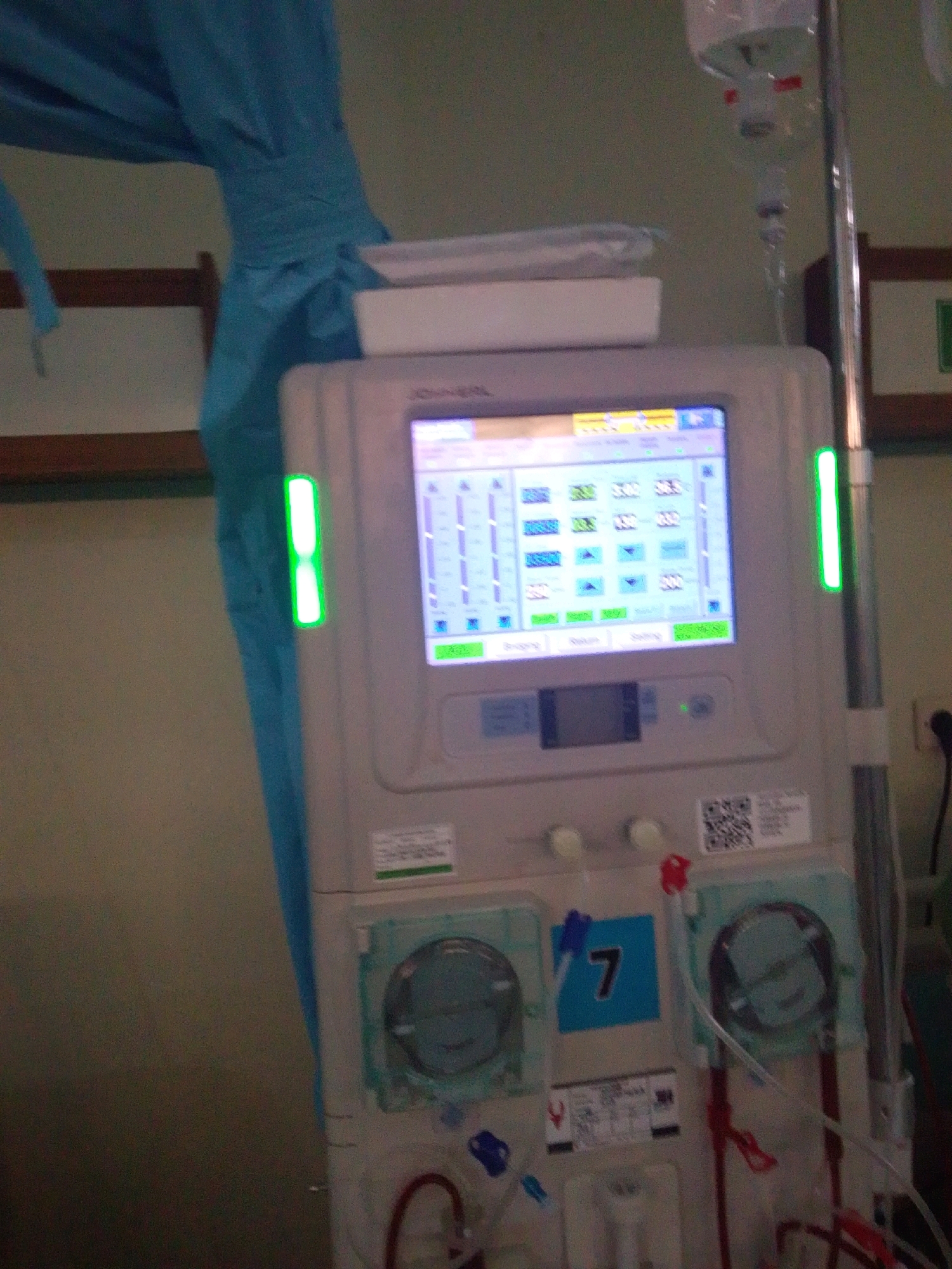 A dialysis machine is shown in a dimly lit room. The machine is white with a touchscreen display showing medical data. There are green indicator lights on the side of the machine, and a blue cloth is draped over the top. Medical tubing and a drip bag are connected to the machine. A blue square with the number "7" is visible on the front of the machine. The background is a plain wall. The scene appears to be in a medical setting, possibly a hospital or clinic in Purworejo, Indonesia, indicated by the presence of medical equipment associated with dialysis.