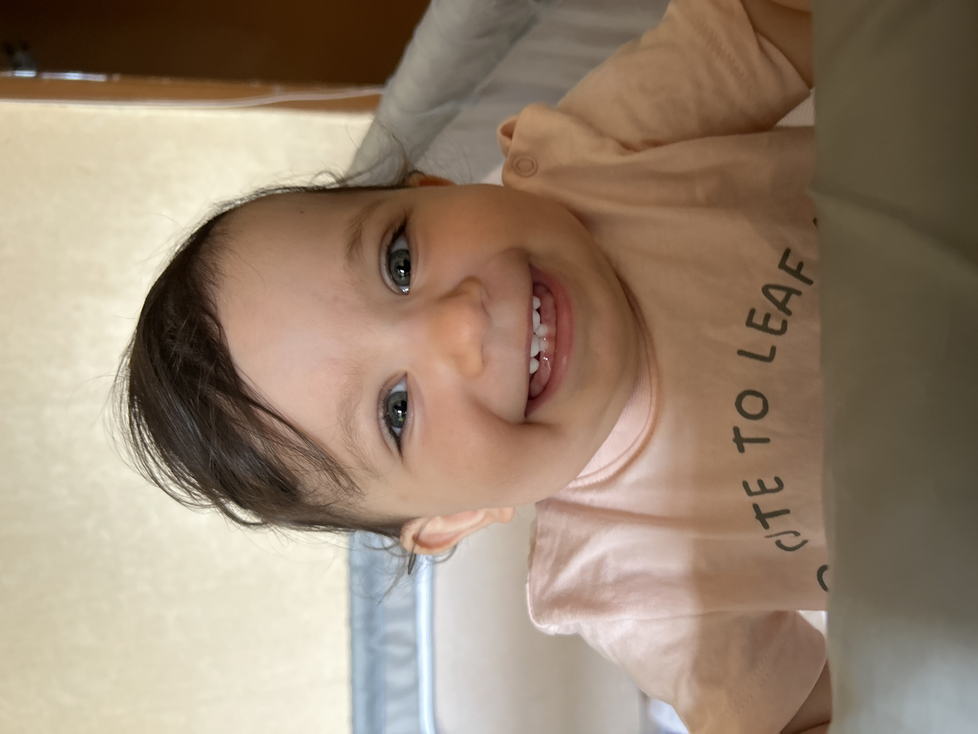 This media file features a close-up portrait of a cheerful baby, likely an infant or young toddler, captured indoors. The baby is looking directly at the camera with a wide, joyful smile, revealing several small, emerging teeth. They have dark, fine hair and striking light-colored eyes, possibly blue or green.
The child is wearing a light peach or salmon-colored long-sleeved top or bodysuit. Visible text on the garment reads "UTE TO LEAF", suggesting the full phrase might be "CUTE TO LEAF" or a similar saying.
The background is softly blurred, indicating a shallow depth of field that keeps the focus on the baby. A light, warm-toned surface, possibly a blanket, cushion, or bed, is visible to the right of the baby. In the upper part of the frame, a darker, warm-brown surface, likely wood or furniture, can be seen. The lighting appears natural, suggesting it is daytime. The overall emotion conveyed is one of happiness and innocence. Although no specific location cues are visible in the image, the context provided indicates the photo was taken in Sofia, Bulgaria.