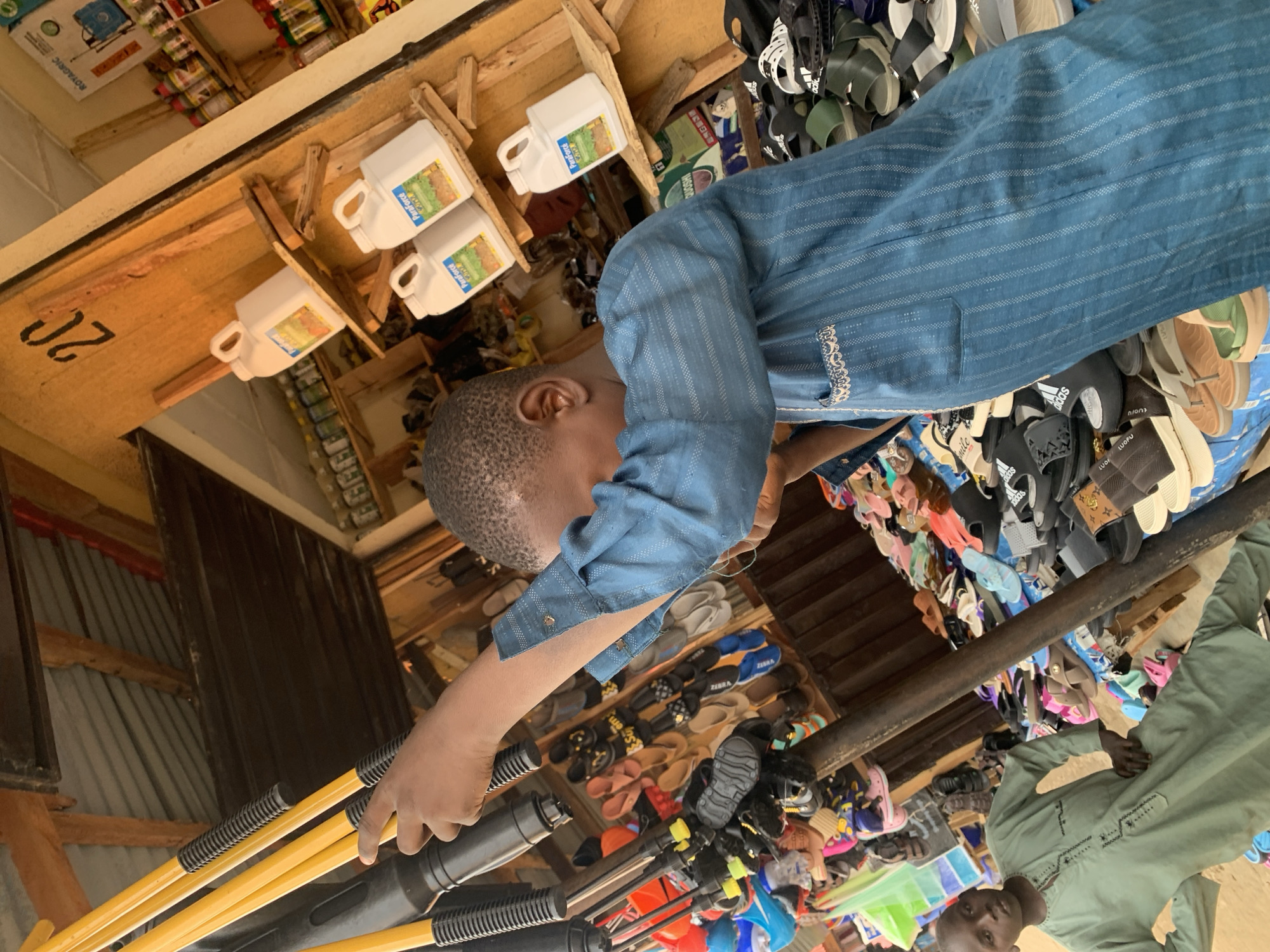 The image captures an indoor scene in what appears to be a small retail store in Kafanchan B, Nigeria. A young person with a shaved head and wearing a blue, striped shirt is seen reaching towards a group of yellow handled implements that are attached to a larger dark piece of equipment. The store's inventory is varied and densely arranged; rows of flip-flops and sandals line one area. Shelves overhead display assorted items, including white bottles labeled with "ParaForce" and "ROYAGRIC." Another individual wearing a green outfit can be seen standing to the right side of the image. The scene provides a glimpse into the local commerce and shopping environment in this Nigerian city.