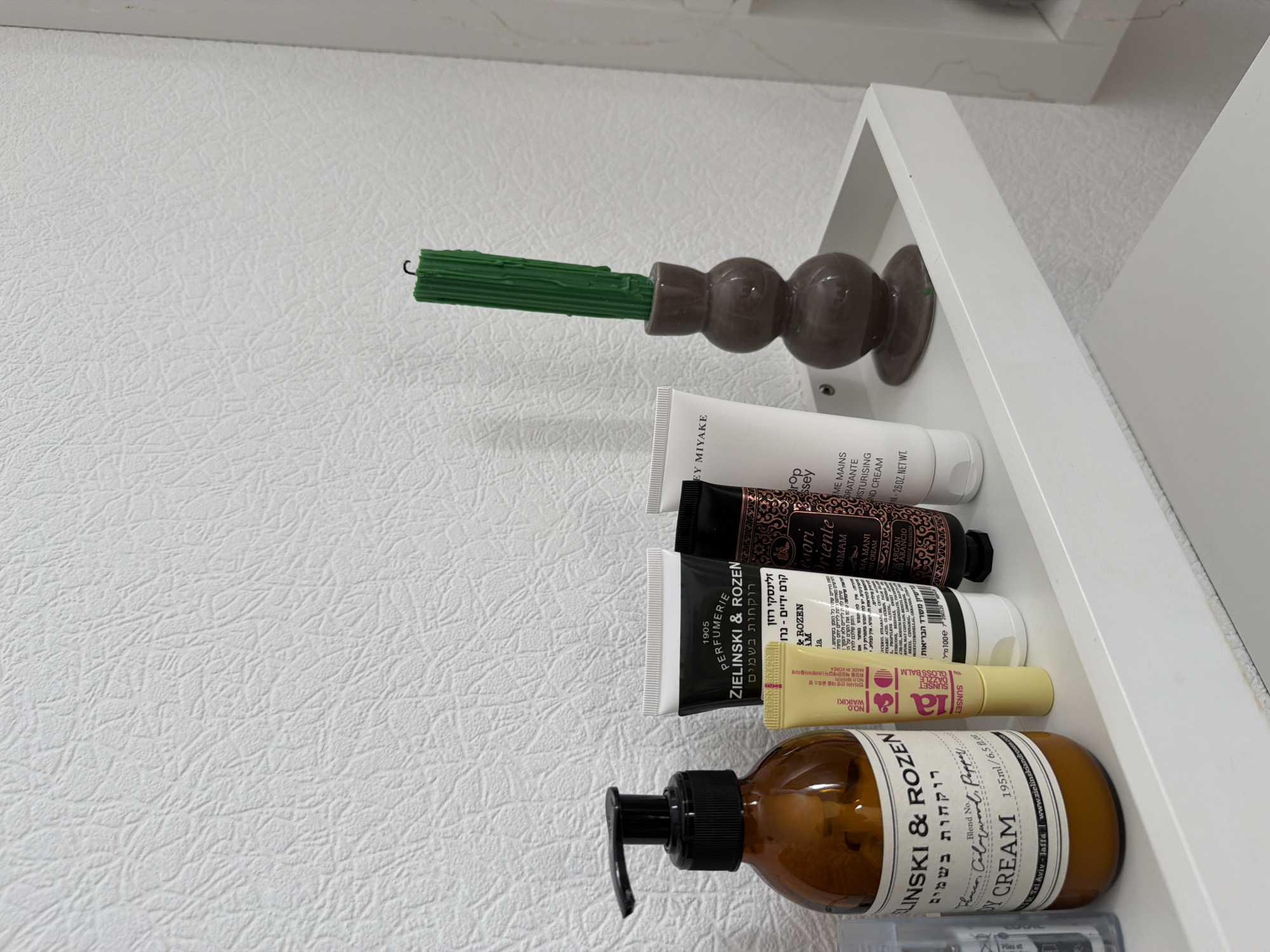 The image shows a white textured wall behind a white shelf. On the shelf, from left to right, are several cosmetic products and a candle holder with a green candle. The products include a large amber bottle with a black pump dispenser, a small yellow tube, a black and white tube, a black tube with ornate gold detailing, and a white tube. A dark brown, spherical candle holder with a fluted candle is positioned to the right of the cosmetic tubes. The overall scene is clean and appears to be an indoor domestic setting. There is no indication of the city of Kyiv, Ukraine, or any specific location.