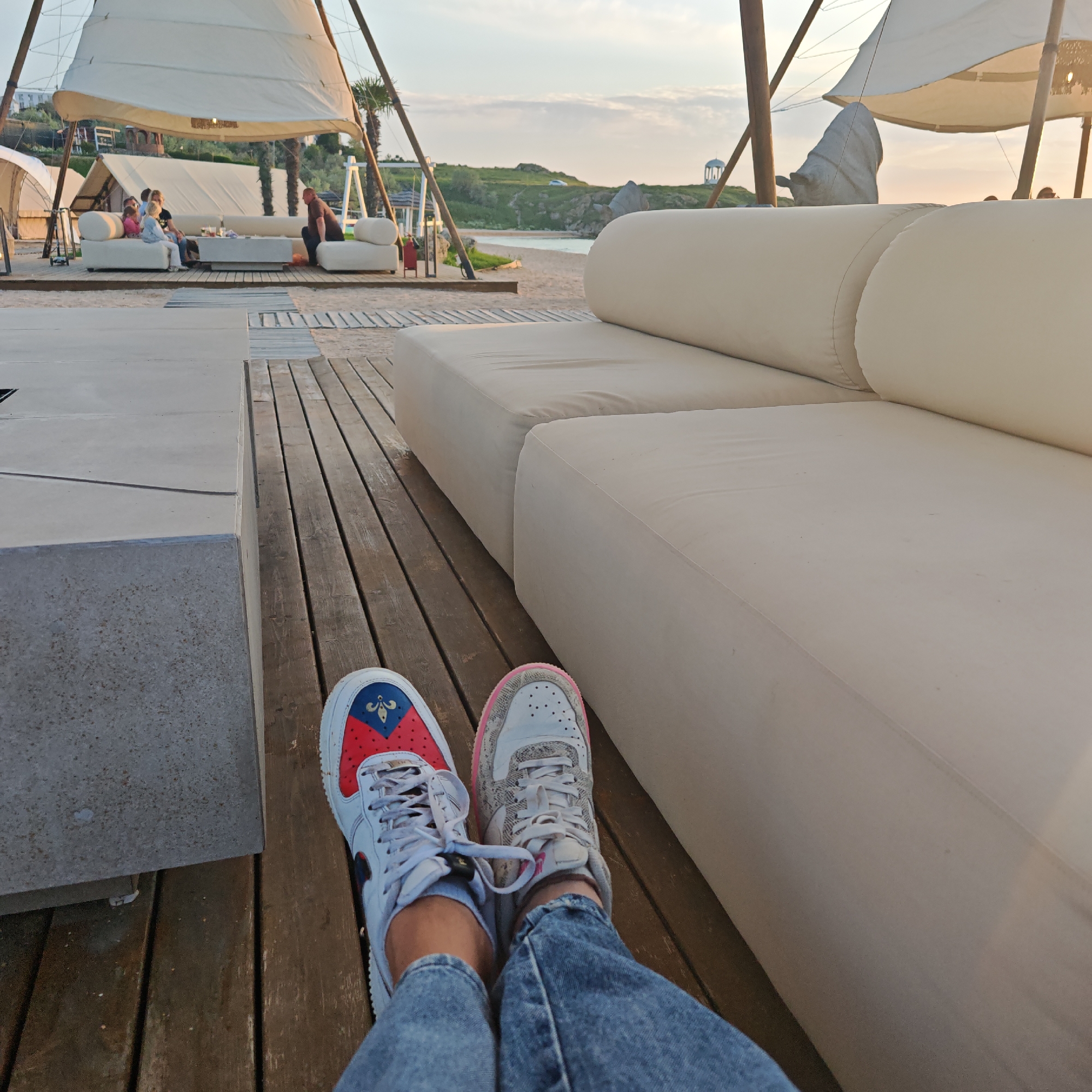 The image shows a first-person view of a person lounging outdoors on a wooden deck in городское поселение Щелкино, Ukraine. They are wearing two different Nike Air Force 1 sneakers and blue jeans. To the left is a light-colored concrete structure. The person is sitting next to a large, white outdoor sofa. In the background, there are several white canopies or tents and people are sitting on sofas. Further in the background, there is a beach area. The lighting suggests it is either early morning or late afternoon.