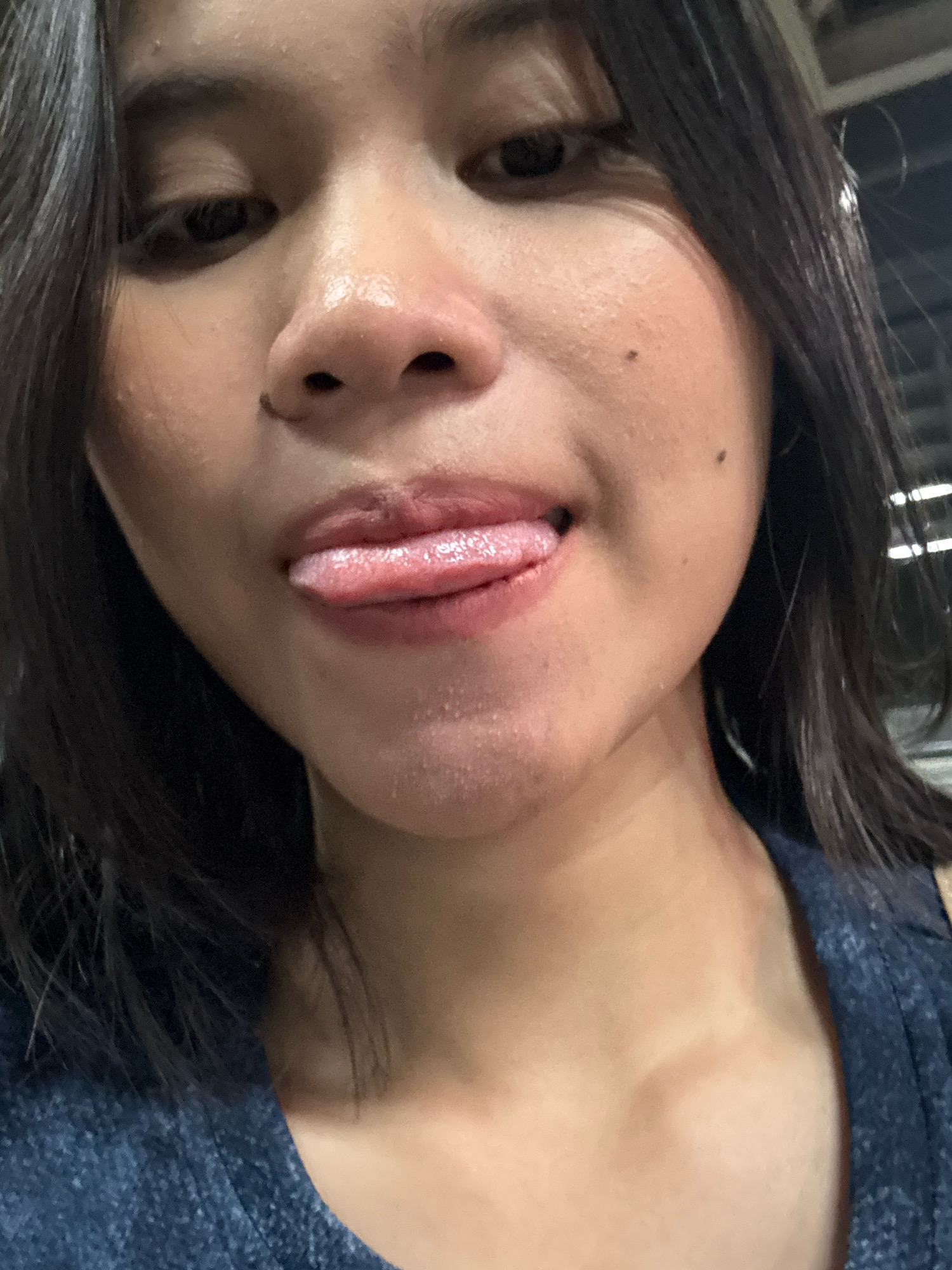 The image is a close-up portrait of a young woman in East Jakarta, Indonesia. Her face is the primary focus, with her tongue playfully extended, touching her lips. She has dark hair and appears to be wearing a dark blue top. The background is blurred, suggesting an indoor or dimly lit environment. The lighting highlights the moisture on her skin and lips, indicating a close-up perspective.