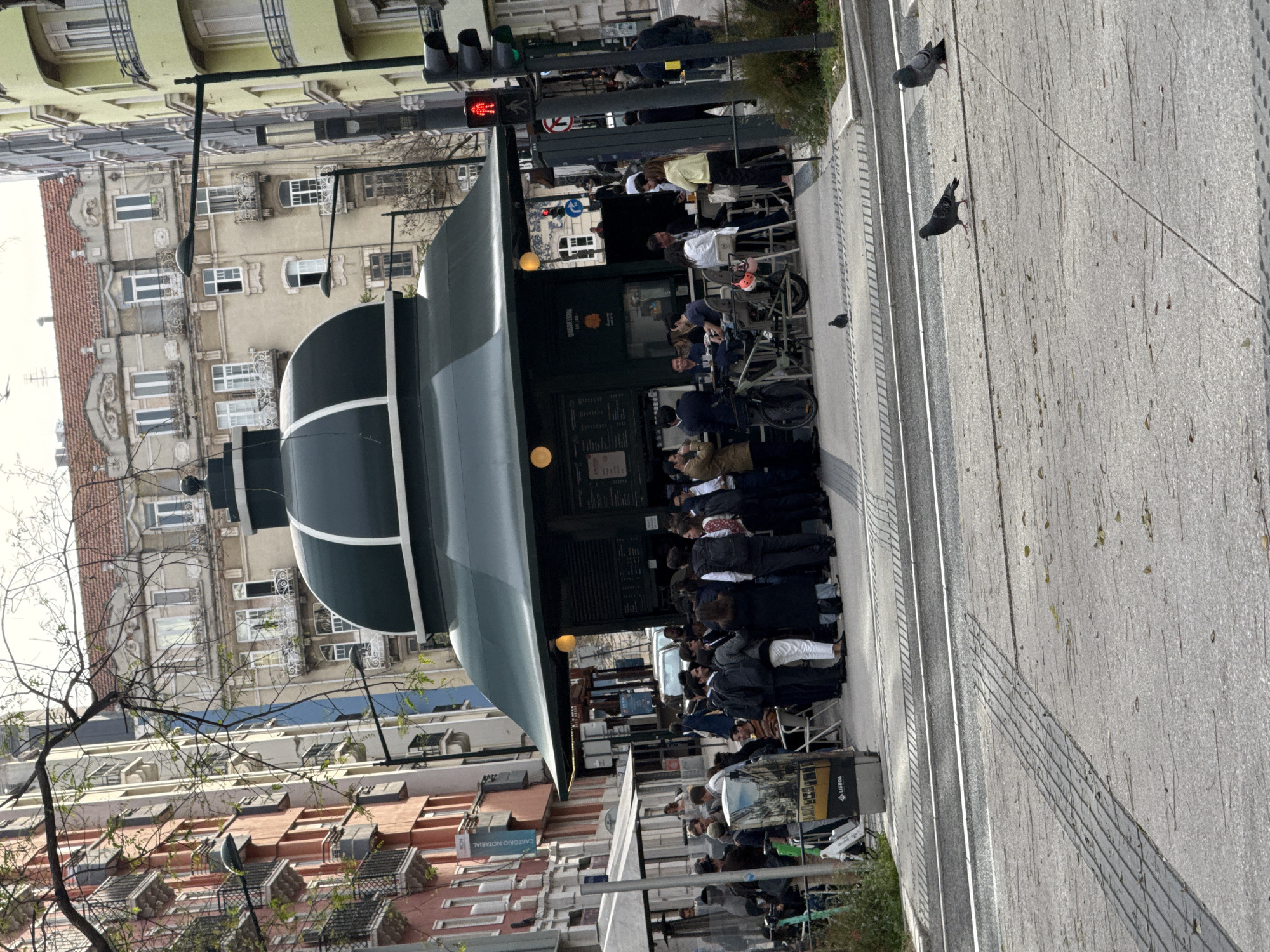 This image captures a vibrant street scene, likely in Lisbon, Portugal, on a bright, sunny day. The focal point is a distinctive dark green kiosk with a multi-tiered, rounded roof, suggesting a traditional city cafe or snack bar. Numerous people are gathered around and near the kiosk; some are standing in line or conversing, while others are seated at outdoor tables and chairs, enjoying food and beverages. Bicycles are parked nearby.
In the background, multi-story buildings with classic European architecture rise, featuring balconies, a variety of window designs, and facades in pale yellow, beige, and terracotta hues. Some buildings have traditional red-tiled roofs. A bare-branched tree partially obscures the view of the buildings on the left.
The foreground and right side of the image show a wide, paved pedestrian area or sidewalk. Several pigeons are scattered on the ground. A red pedestrian traffic light and a green vehicle traffic light are visible in the upper left, indicating a street intersection. The clear sky and strong shadows suggest it is daytime, possibly mid-morning or early afternoon.
Visible text on the kiosk reads "QUIOSQUE LISBOA," and the word "LISBOA" is also seen on a grey stand near the outdoor seating, confirming the city. The overall scene depicts a lively urban environment, characteristic of a European capital.