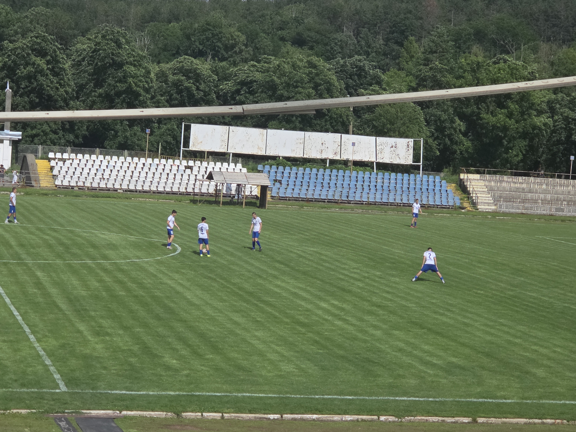 An aerial view captures a grassy soccer field at a stadium in Targovishte, Bulgaria, on a sunny day. Several young men, wearing white jerseys with blue shorts and varying numbers on their backs, are scattered across the field. Some are standing, while one is in a wide-legged stretching pose. The field is marked with white lines, indicating boundaries and play areas. Behind the field, rows of empty stadium seats, some white and some blue, are visible. Further back, a dense line of lush green trees forms a natural backdrop against the sky. The overall scene depicts a training or practice session on a well-maintained sports ground.