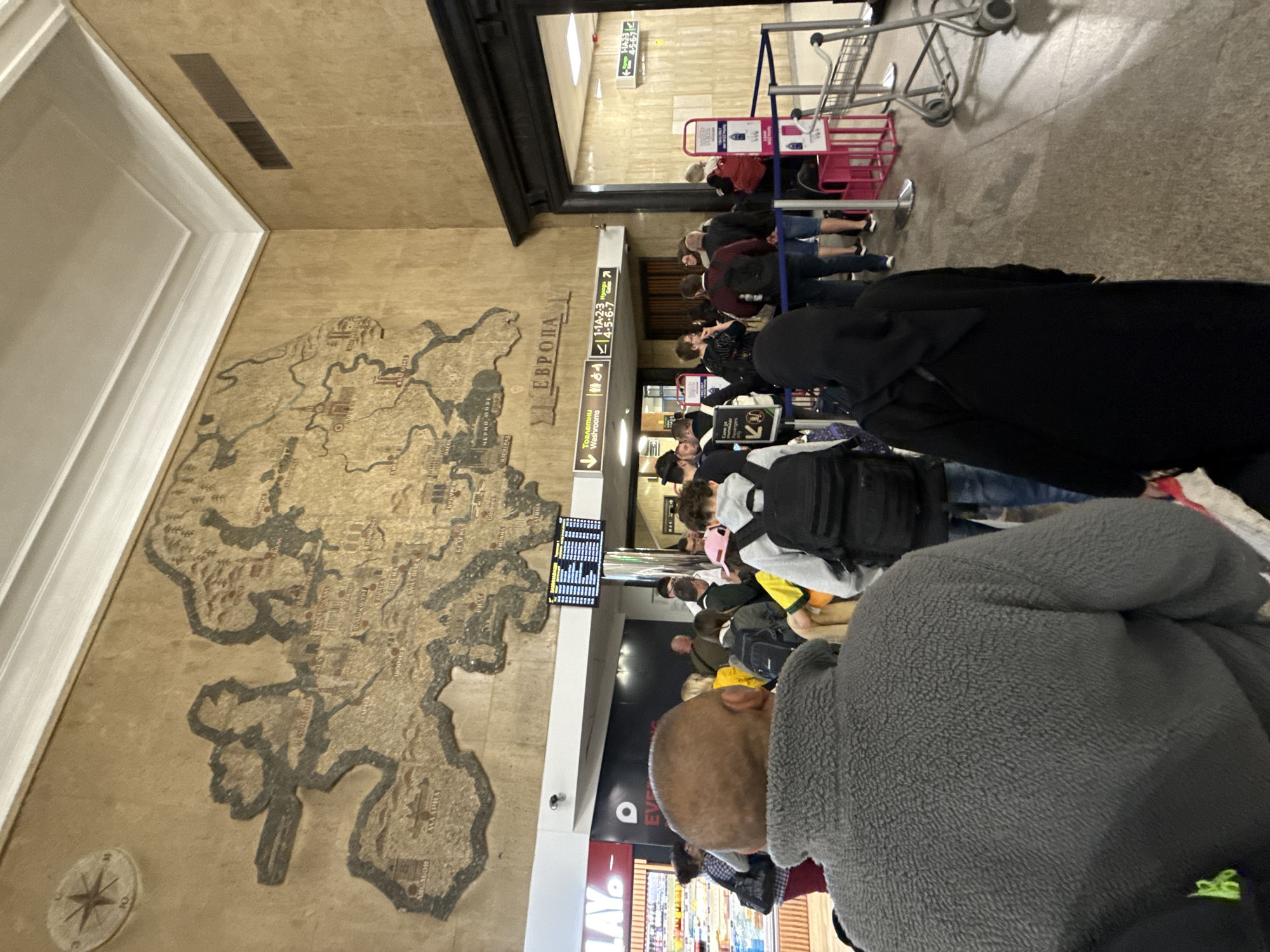 This image captures an indoor scene, likely a bustling transportation hub such as a train or bus station in Sofia, Bulgaria. A long queue of people, primarily adults and young adults, stretches into the distance, with many individuals carrying backpacks and luggage, indicating they are travelers. The queue is organized by blue and white stanchions, and several empty metal luggage carts are positioned further down the line.
A dominant feature of the setting is a large, elaborate mosaic map displayed on a light brown wall to the left. This artistic map illustrates Europe, with numerous cities and regions labeled in Cyrillic script. Prominently visible texts include "ЕВРОПА" (EUROPA), "БЪЛГАРИЯ" (BULGARIA), "СОФИЯ" (SOFIA), "ПЛОВДИВ" (PLOVDIV), "ЧЕРНО МОРЕ" (BLACK SEA), "ВАРНА" (VARNA), "БУРГАС" (BURGAS), "РУСЕ" (RUSE), "ИСТАНБУЛ" (ISTANBUL), and "АТИНА" (ATHENS), confirming the map's focus on the Balkans and Eastern Europe. A compass rose is also present on the wall beneath the map.
Overhead, a digital display board shows columns of information, likely schedules or announcements, though the specific text is unreadable due to blurriness. Below it, a white hanging sign provides directions, featuring "Тоалетни" (Toilets) and "Washrooms" with a directional arrow, alongside symbols for male, female, and disabled access. Other visible text on this sign includes "1-14-203" and "1-4-5-6-7," which likely refer to platform or gate numbers, and "Изход" (Exit). The overall lighting is artificial, typical of an indoor public facility. Individuals in the queue are dressed casually, and while their faces are mostly obscured, they appear to be waiting calmly. Notable subjects in the foreground include a person wearing a grey fleece jacket and, further back, an individual in a black abaya or cloak.