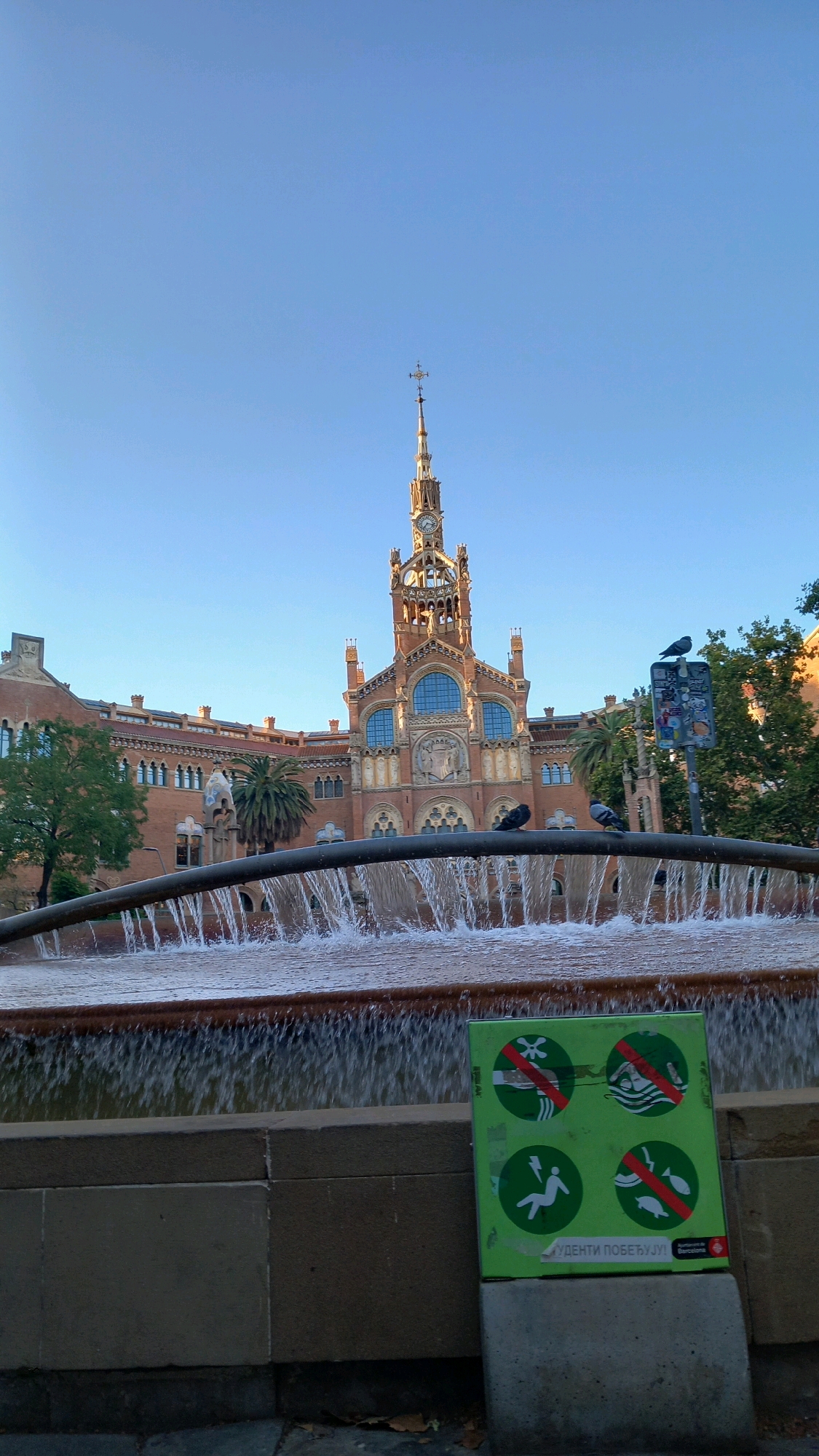 The image captures the Recinte Modernista de Sant Pau in Barcelona, Spain, under a clear blue sky. In the foreground, a stone structure supports a fountain with water cascading over its edge, creating a foamy texture. A dark, curved pipe runs across the middle of the frame, also draped with falling water.
On the lower right, a green sign with four circular red-and-white prohibition symbols is prominently displayed. These symbols depict actions such as diving, swimming, and touching aquatic animals. Below the symbols, text in a non-Latin script is visible, along with a logo that reads "Ajuntament de Barcelona."
In the mid-ground, a large, ornate building with brick facade and numerous arched windows stands behind the fountain. A central tower with a clock and a cross on top dominates the building's architecture. Several pigeons are perched on a railing in front of the building and on a signpost to the right of the tower. Trees, including a palm tree, are visible around the building, suggesting an outdoor setting with landscaping. The overall scene conveys a sense of architectural grandeur and public space.