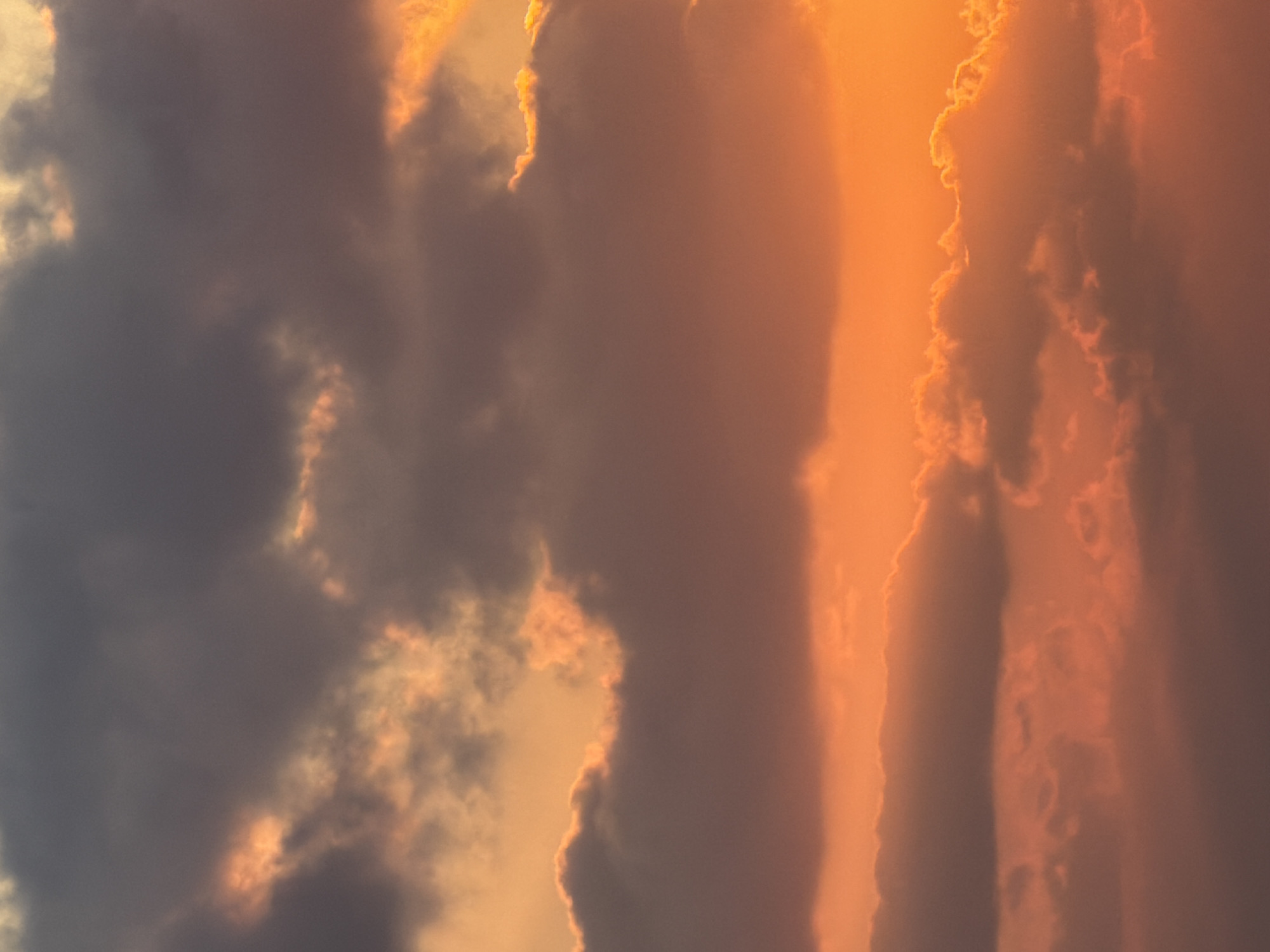 The image is a close-up, vertical, outdoor photograph of the sky during sunset. The sky is filled with clouds, ranging in color from dark gray to vibrant orange and yellow. The sunlight is illuminating the edges and undersides of the clouds, creating a dramatic and colorful effect. There are no discernable landmarks or structures that would indicate a specific location within Tel Aviv, Israel. The scene is purely atmospheric and abstract.