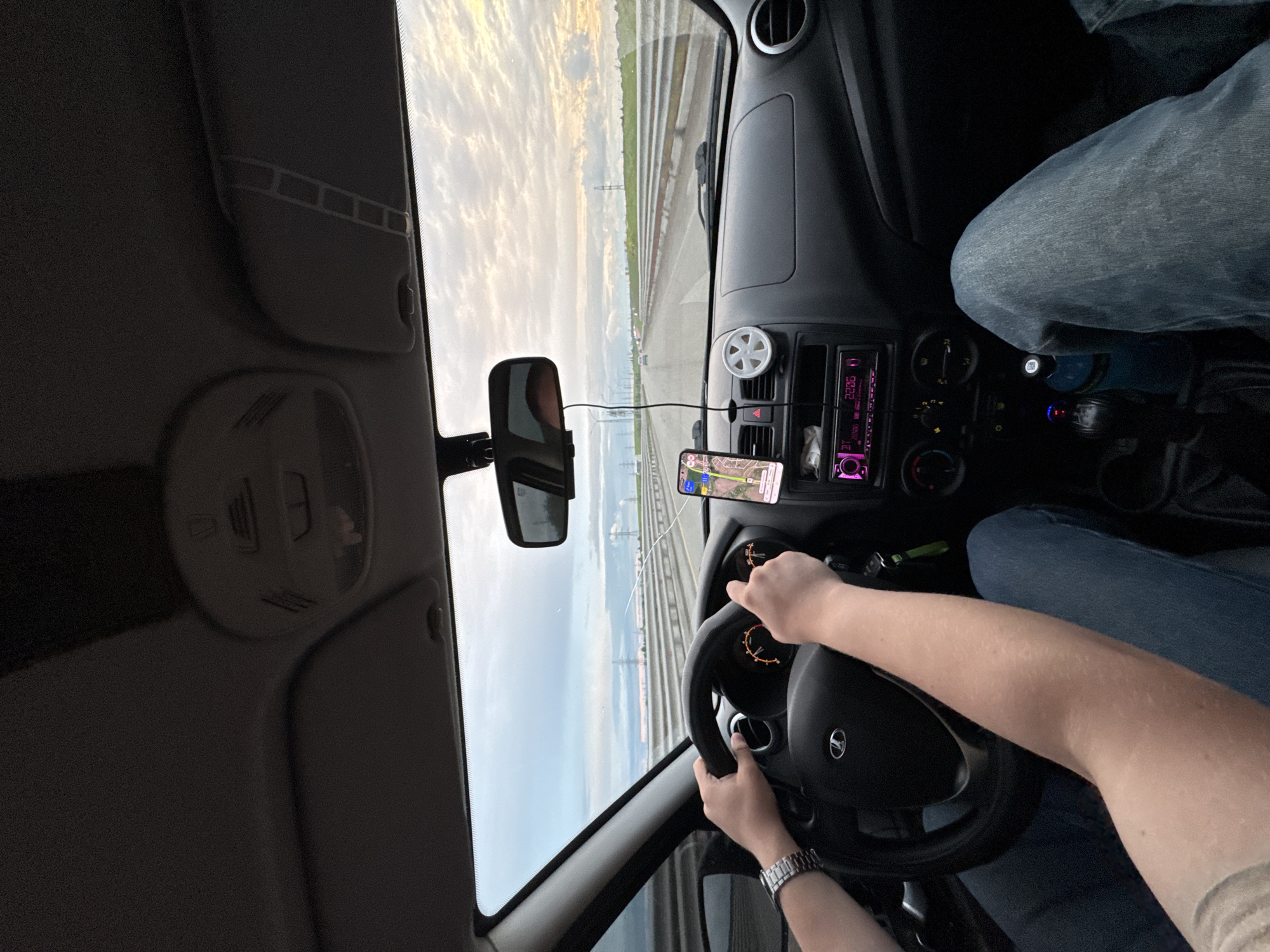 The image captures the interior of a car moving on a highway, likely taken by a passenger. The driver's hands are visible on the steering wheel, wearing a watch on their left wrist. A smartphone is mounted on the dashboard, displaying a map application. The dashboard features a radio and various controls. Outside the car, the landscape shows a highway with parallel lines and structures resembling electrical pylons under a sky with scattered clouds. Considering the query, the location is approximated to be Bol'shaya Mos', Russia, but this is inferred from the user's query and cannot be definitively confirmed from the image alone. There are no interactions visible between people; the primary action is driving. The overall environment is a car interior and a highway landscape.