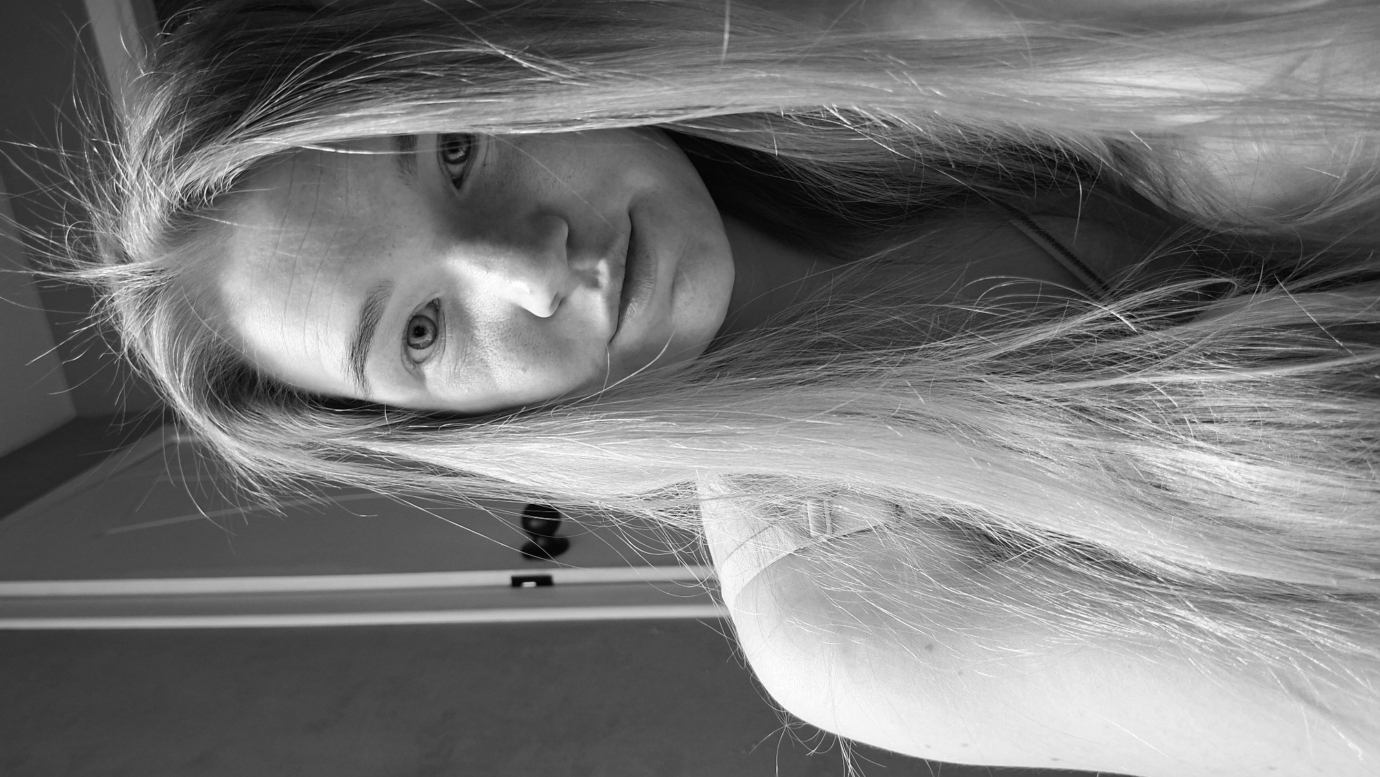 A grayscale, upside-down, close-up shot of a fair-skinned person with long, straight, light-colored hair framing their face. The person has freckles, light eyes, and a neutral expression. Part of their shoulder and upper arm is visible in the lower right corner of the image. The background shows part of a wall, a door frame, and the door. There is no indication of the location in the city of Chaikovsky, Russia.