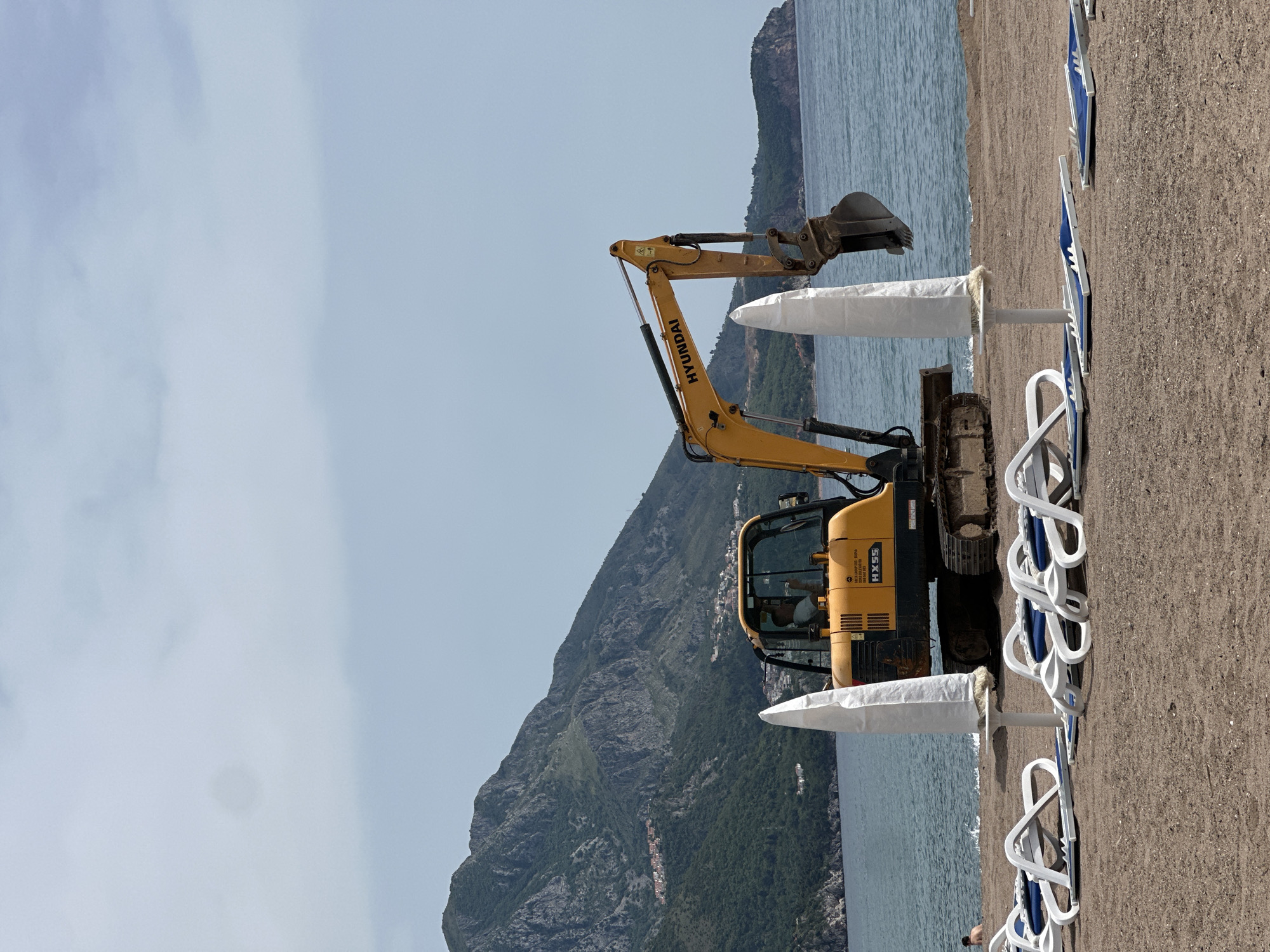 A yellow Hyundai excavator sits on a sandy beach in Boreti, Montenegro. The excavator is next to closed, white beach umbrellas and blue and white lounge chairs. In the background, there is a mountain with some structures on it, and the sea. The sky is overcast.