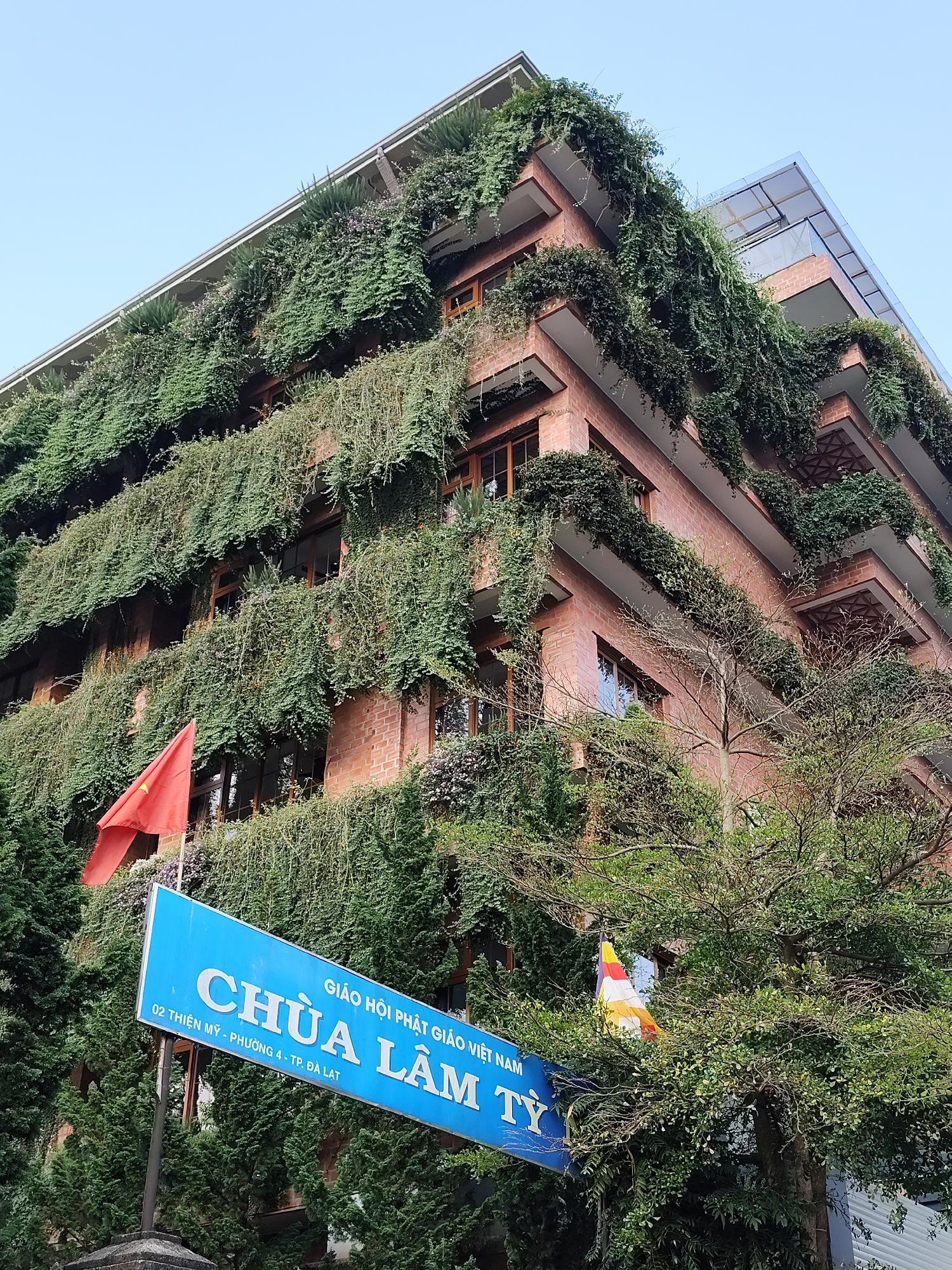 The image depicts a multi-story brick building covered in lush green vegetation, located in Da Lat, Vietnam. The building appears to be a Buddhist temple ("Chùa" in Vietnamese), as indicated by the blue sign with the text "CHÙA LÂM TỪ GIÁO HỘI PHẬT GIÁO VIỆT NAM" and an address, along with a Buddhist flag partially visible behind a tree. A Vietnamese flag is also visible, attached to the building. The building's architecture features balconies and a partially glass roof. The abundant greenery drapes over the building's facade, creating a "living wall" effect. The sky is light blue. Trees surround the building, obscuring the view of the ground.