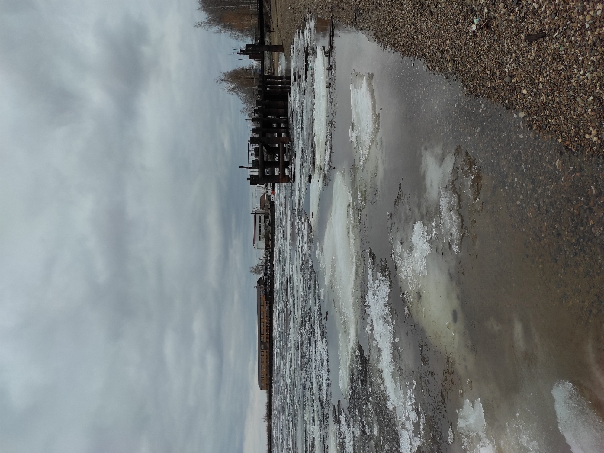 This media file captures a bleak, overcast winter or early spring scene along a body of water in Chaikovsky, Russia. The image is dominated by a grey, cloudy sky that stretches across the top.
In the foreground and midground, a significant portion of the frame is filled with water, partially covered by large, broken sheets and chunks of ice. The water's surface reflects the dull sky, creating a muted palette. To the right, a rocky or gravelly shoreline borders the water. The pebbles are varied in size and color, mostly browns and greys.
Along the left edge of the water, several weathered, dark wooden pilings or remnants of a pier or dock stand out against the grey water and sky. Beyond these structures, a line of bare trees suggests the cold season. Further in the distance, indistinct buildings with flat roofs are visible, indicating an urban or industrial area along the waterfront.
The overall impression is one of cold, stillness, and the transition of seasons, with the ice breaking up. There are no visible people or explicit activities taking place, and no discernable text. The time of day appears to be daytime, under heavily overcast conditions.
