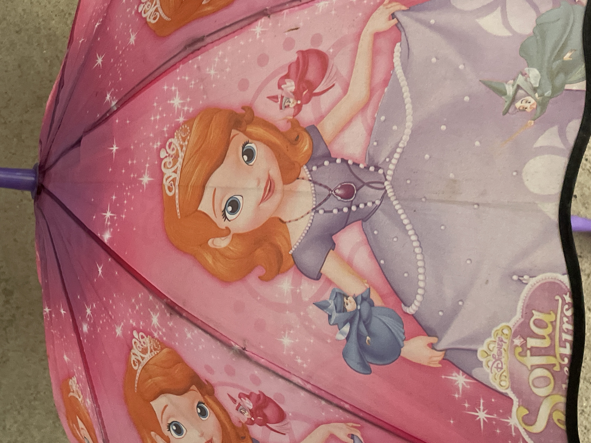 The image displays a close-up of a pink and purple umbrella featuring artwork of Disney's Sofia the First. Sofia is depicted in a purple dress with a pearl necklace and a tiara. She is interacting with two fairy characters. One fairy, in a blue outfit, is perched on her arm, while another fairy in a green witch's hat is flying near the hem of her dress, holding a wand. The background of the umbrella is decorated with pink and purple patterns, including stars and polka dots. A logo for "Sofia the First" is visible at the bottom. The umbrella is open and appears to be resting on a textured, grey surface. The image does not provide any information about its location in Tashkent, Uzbekistan.