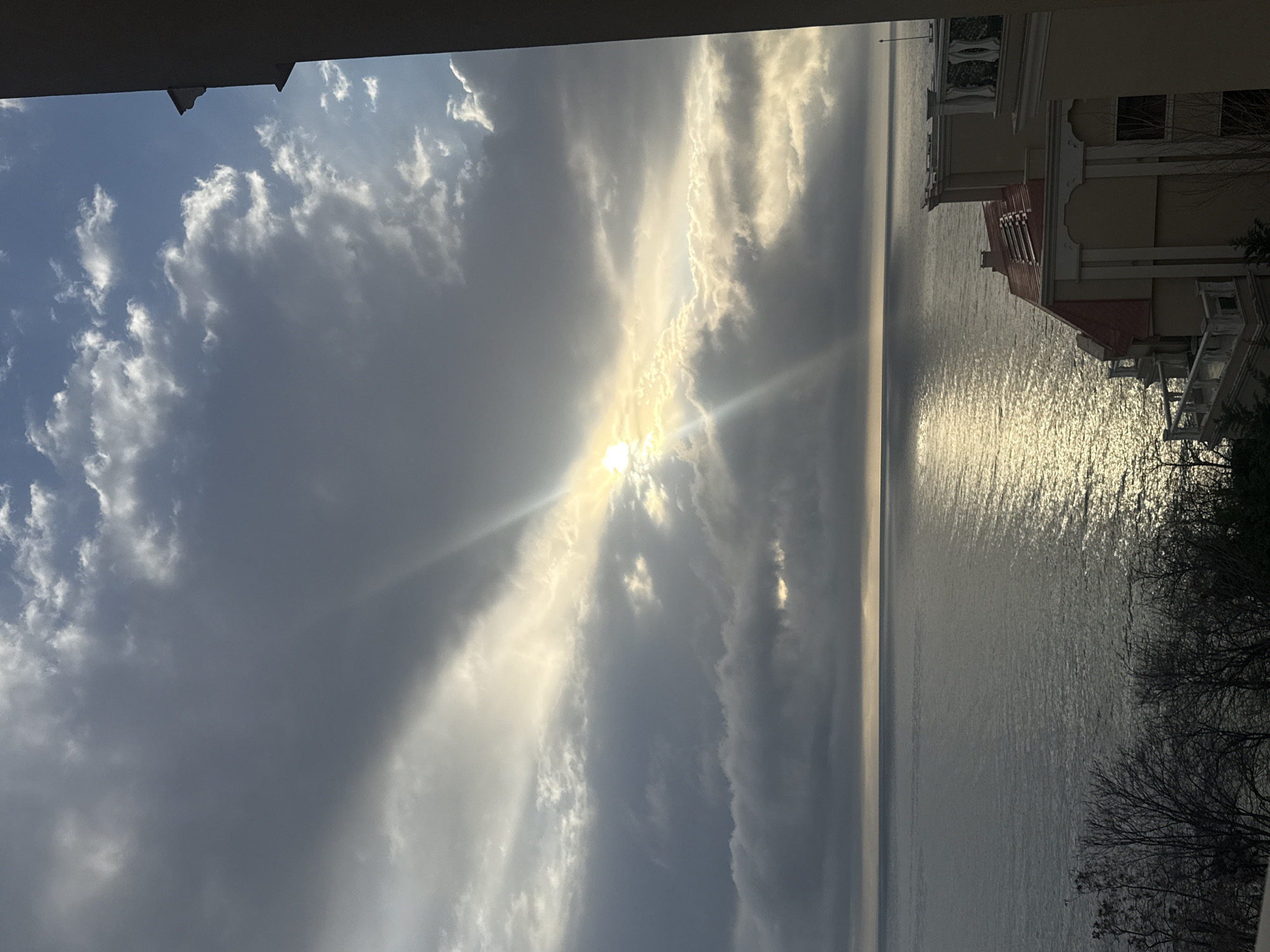 View from a balcony in Varna, Bulgaria, showing a body of water (likely the Black Sea) and a sky with scattered clouds and the sun shining through them. A building is visible on the right side of the frame. Bare tree branches are partially visible on the right edge of the image. The scene is illuminated by daylight.