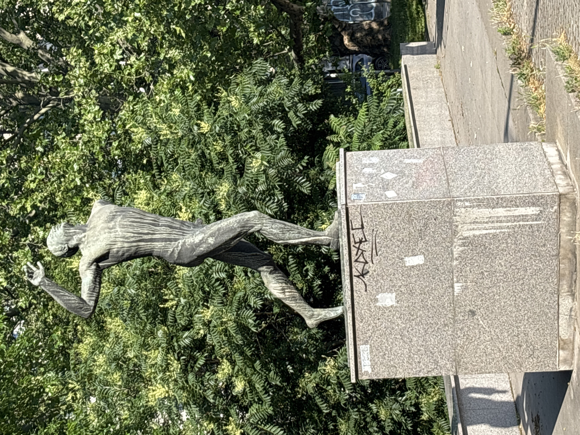 A bronze statue of a male athlete in a running pose stands on a granite pedestal. The statue is weathered and shows streaks of discoloration. The pedestal is also stained and has graffiti tags on its surfaces. The background consists of lush green trees and foliage. The scene appears to be outdoors in a park or public space. The statue is located in Sofia, Bulgaria, at the "National Palace of Culture" (NDK).
