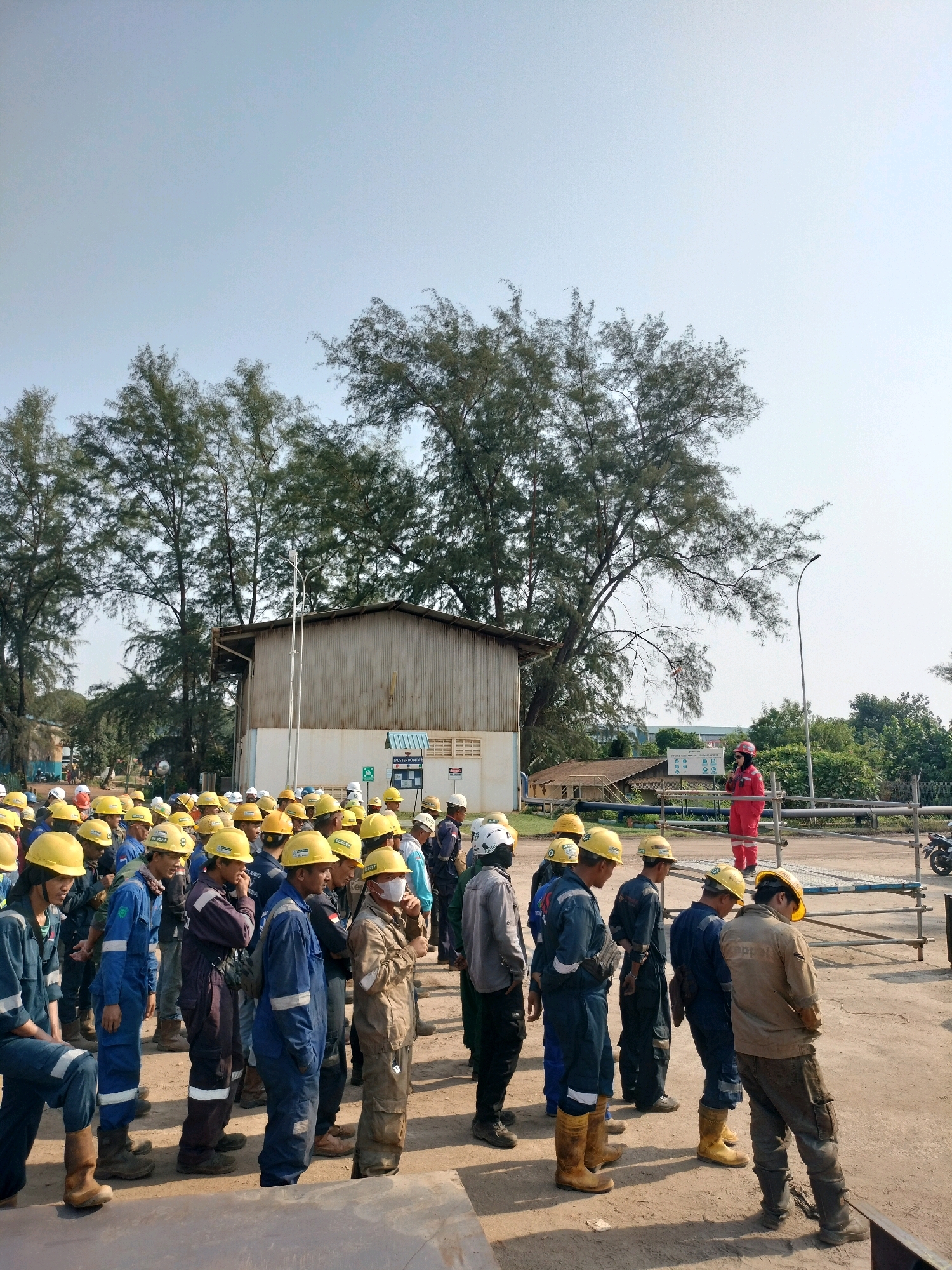 The image depicts a group of workers, mostly wearing yellow hard hats and blue or beige coveralls, gathered outdoors in what appears to be an industrial or construction site. The setting is likely Batam, Indonesia, given the context provided. In the foreground, several workers are standing in rows, some looking towards a person in a red jumpsuit who is standing on a raised platform. This individual in red seems to be addressing the group. Behind the workers, there is a large, corrugated metal building and several trees. The ground is dusty and uneven, with some scaffolding structures visible. The sky is clear and blue. The overall scene suggests a safety briefing or a pre-work gathering.